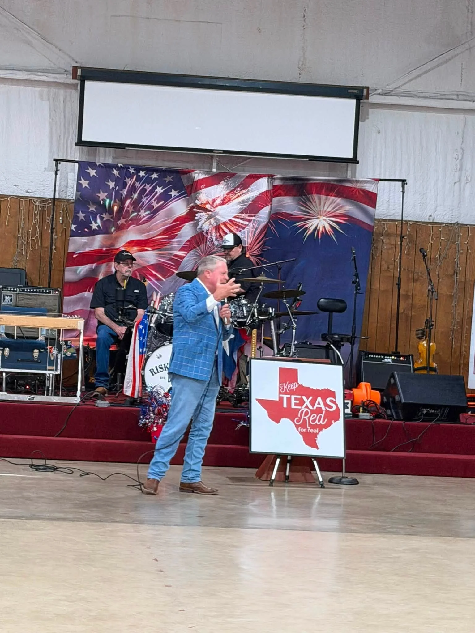 A man in a blue checkered suit speaking on a microphone on a stage decorated with a patriotic Texas and American flag backdrop. Behind him are musicians and equipment, and a sign reads 'Keep Texas Red for real' with an outline of Texas.