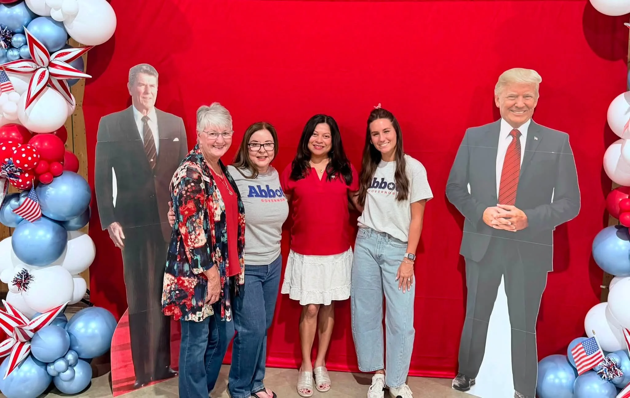 Four women are standing in front of a decorated red background with patriotic balloon arrangements. Two cardboard cutouts of former U.S. Presidents Ronald Reagan and Donald Trump are on either side. The women are smiling and wearing casual clothing, 