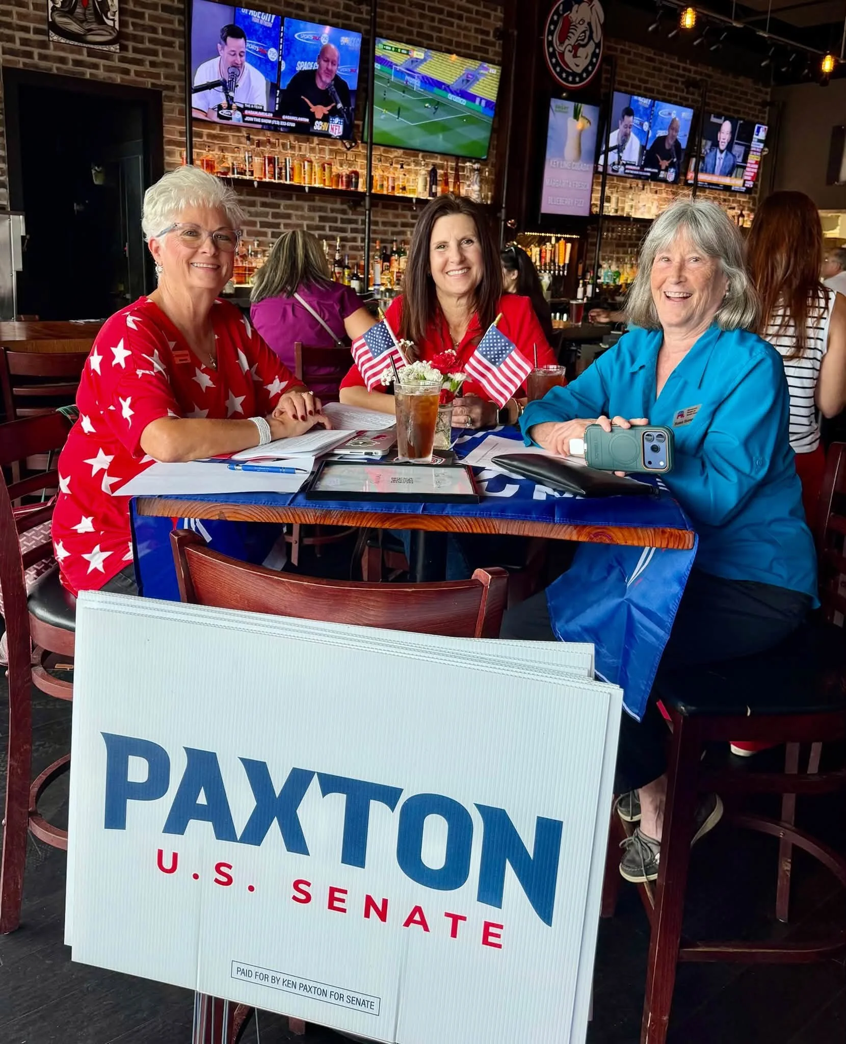 Three women sitting at a table in a bar or restaurant, smiling at the camera, with TV screens showing sports in the background. A large poster with the text 'PAID FOR BY KEN PAXTON FOR SENATE' is in front of them.