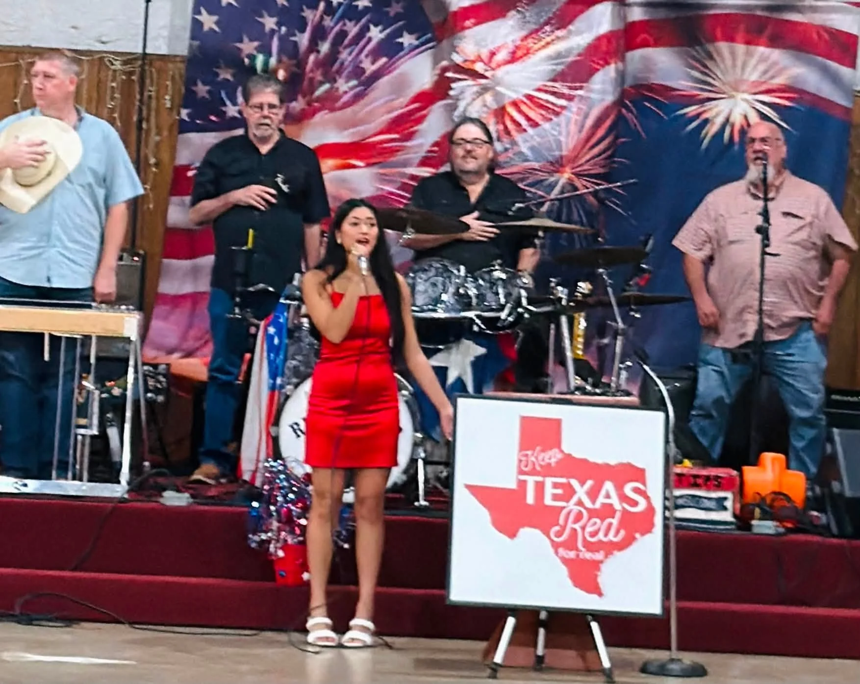 A group of five people on a stage with American flags and fireworks-themed backdrop, including a young woman in a red dress and white shoes singing into a microphone, and four men standing behind her with musical instruments and microphones, one hold