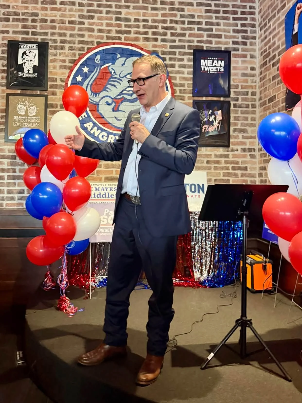 Man in a suit and glasses speaking into a microphone at a political event with red, white, and blue balloons and campaign signs, including a poster for Mayes Middleton for U.S. Senate, in front of a brick wall with political posters and a large logo 