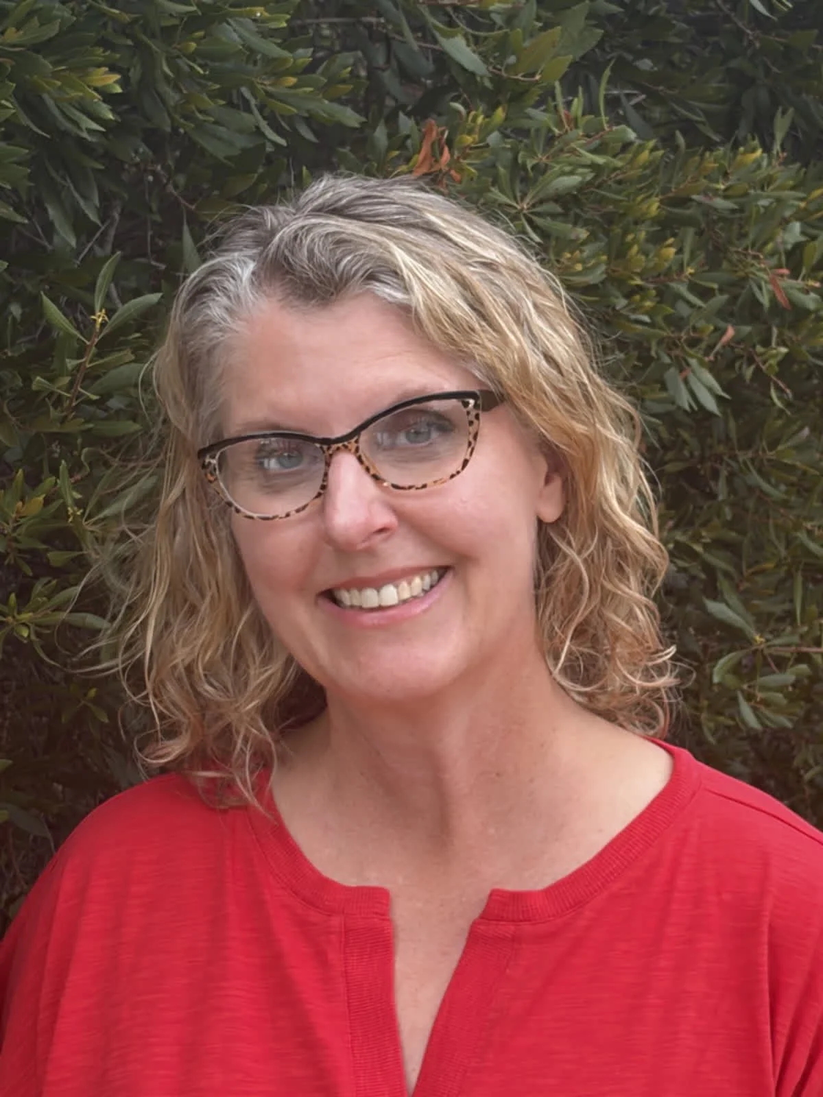 A woman with blonde, curly hair and glasses with a leopard print pattern, smiling and wearing a red top, standing in front of green leafy plants.