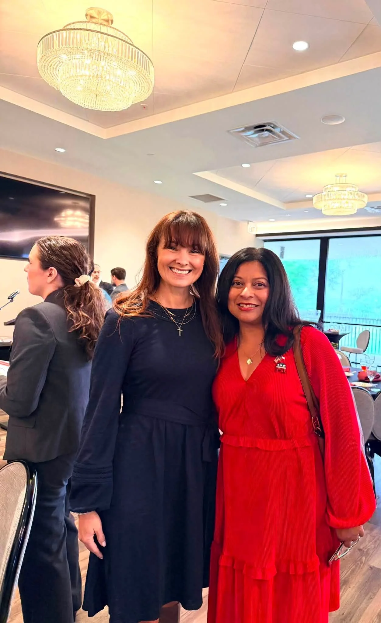 Two women smiling and standing together at an indoor event, one in a navy dress with a cross necklace, the other in a red dress with a pin and a red handbag, with chandeliers and a window in the background.