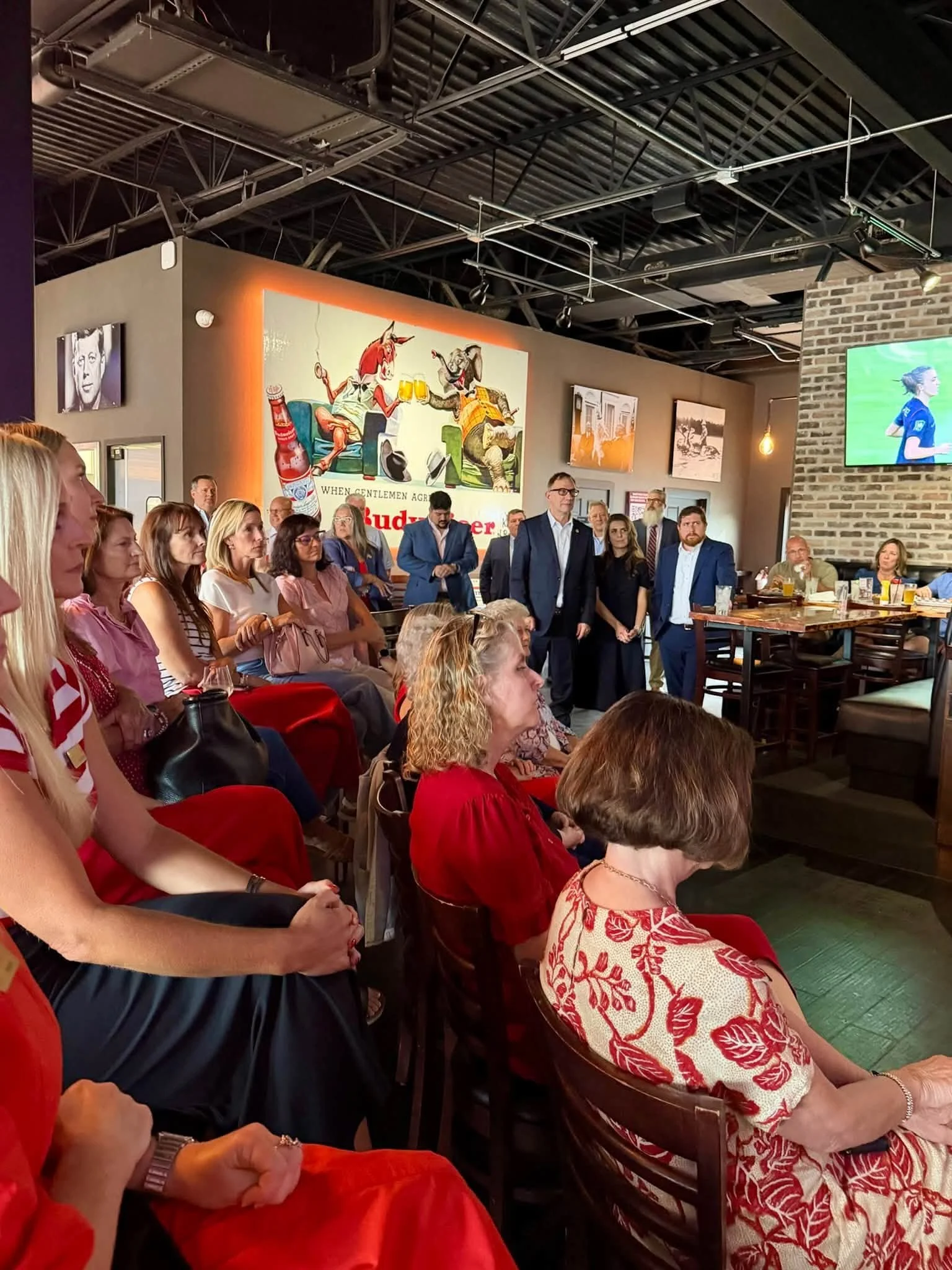 Indoor gathering of people listening to a speaker, with artwork on the walls and a television screen showing a person in a blue outfit, in a restaurant or event space.