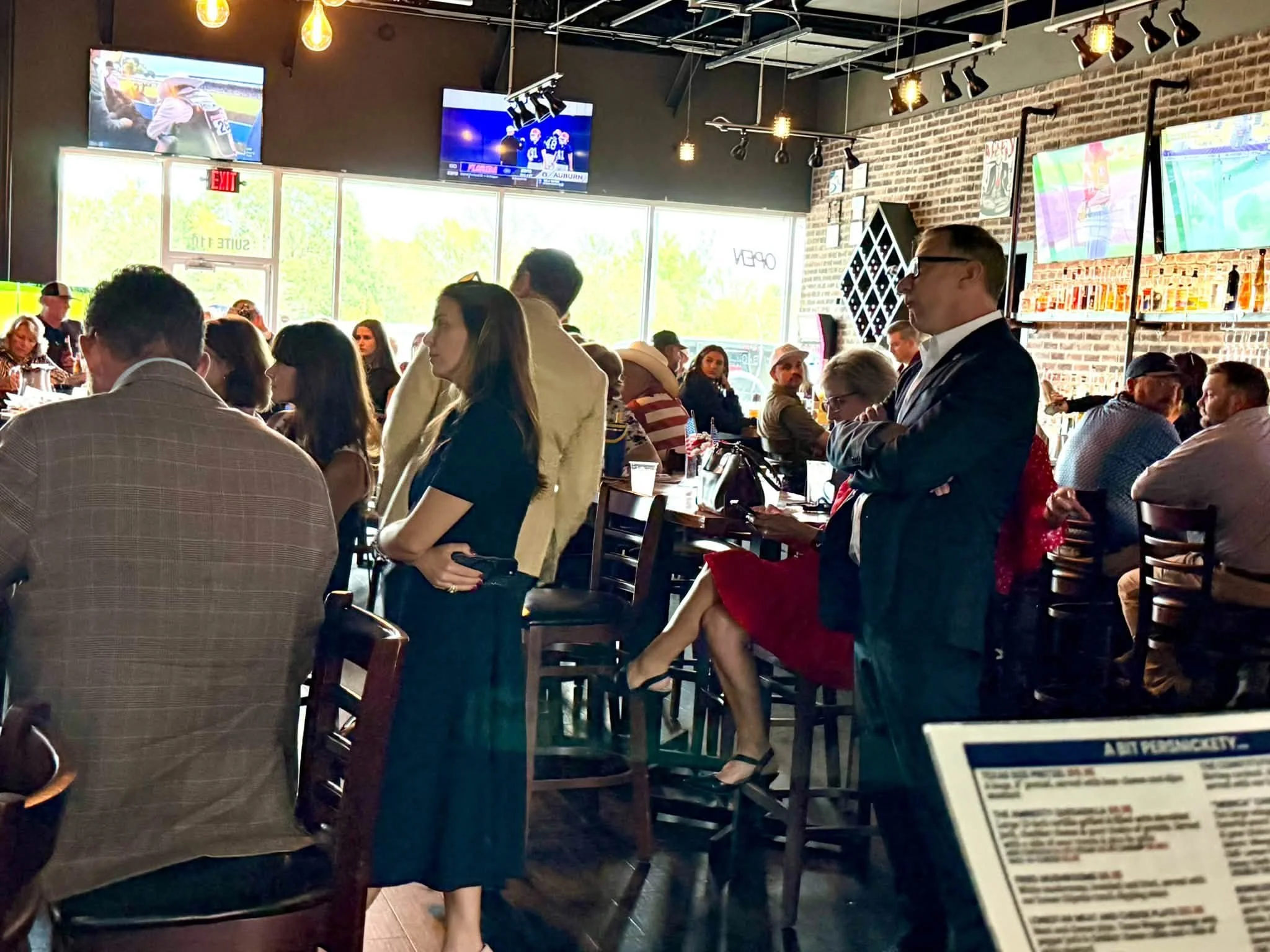 Crowd sitting and standing at a sports bar, watching football games on multiple TVs, with a brick wall and large windows in the background.