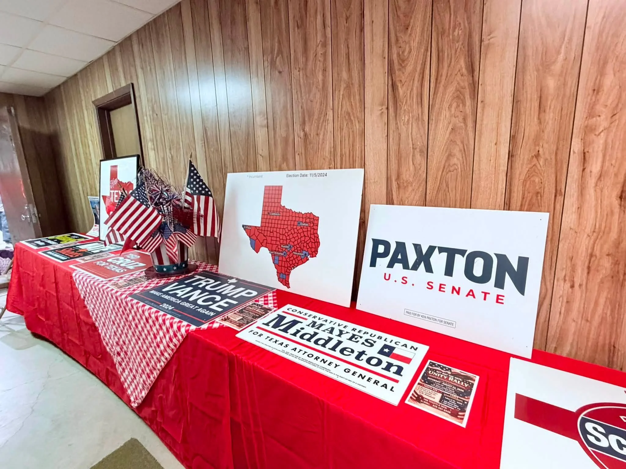 Table covered with political campaign materials including signs, posters, and small American flags, with a wood-paneled wall in the background.