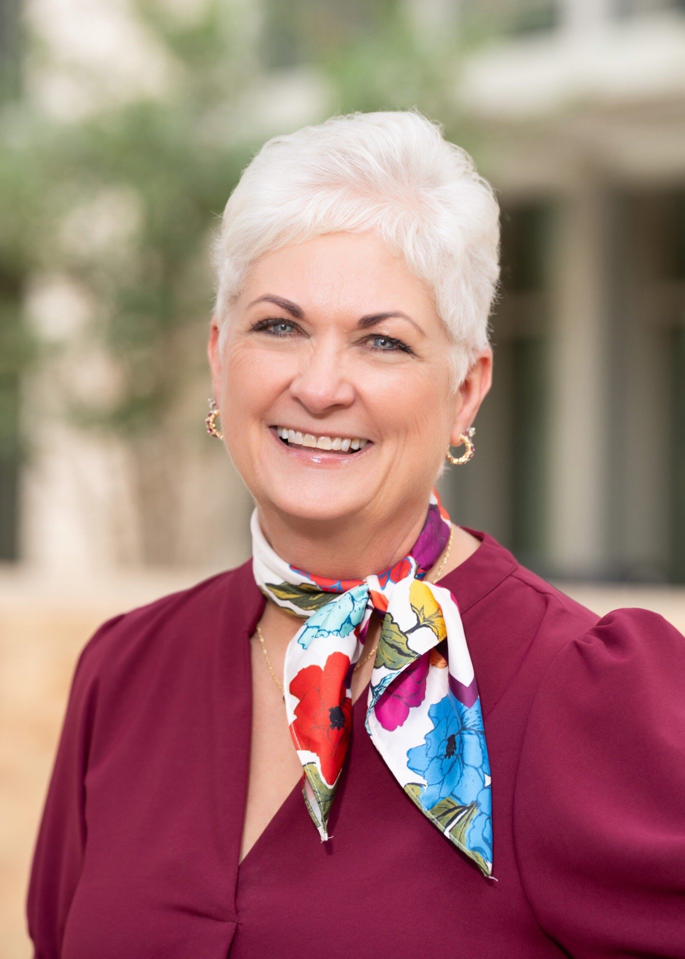 A woman with short white hair smiling outdoors, wearing a maroon blazer and a colorful floral scarf.