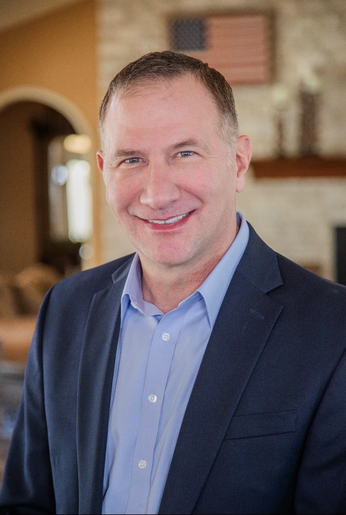 A middle-aged man with short brown hair and blue eyes dressed in a light blue shirt and dark blue blazer, smiling at the camera indoors with a blurred background featuring an American flag and a brick wall.