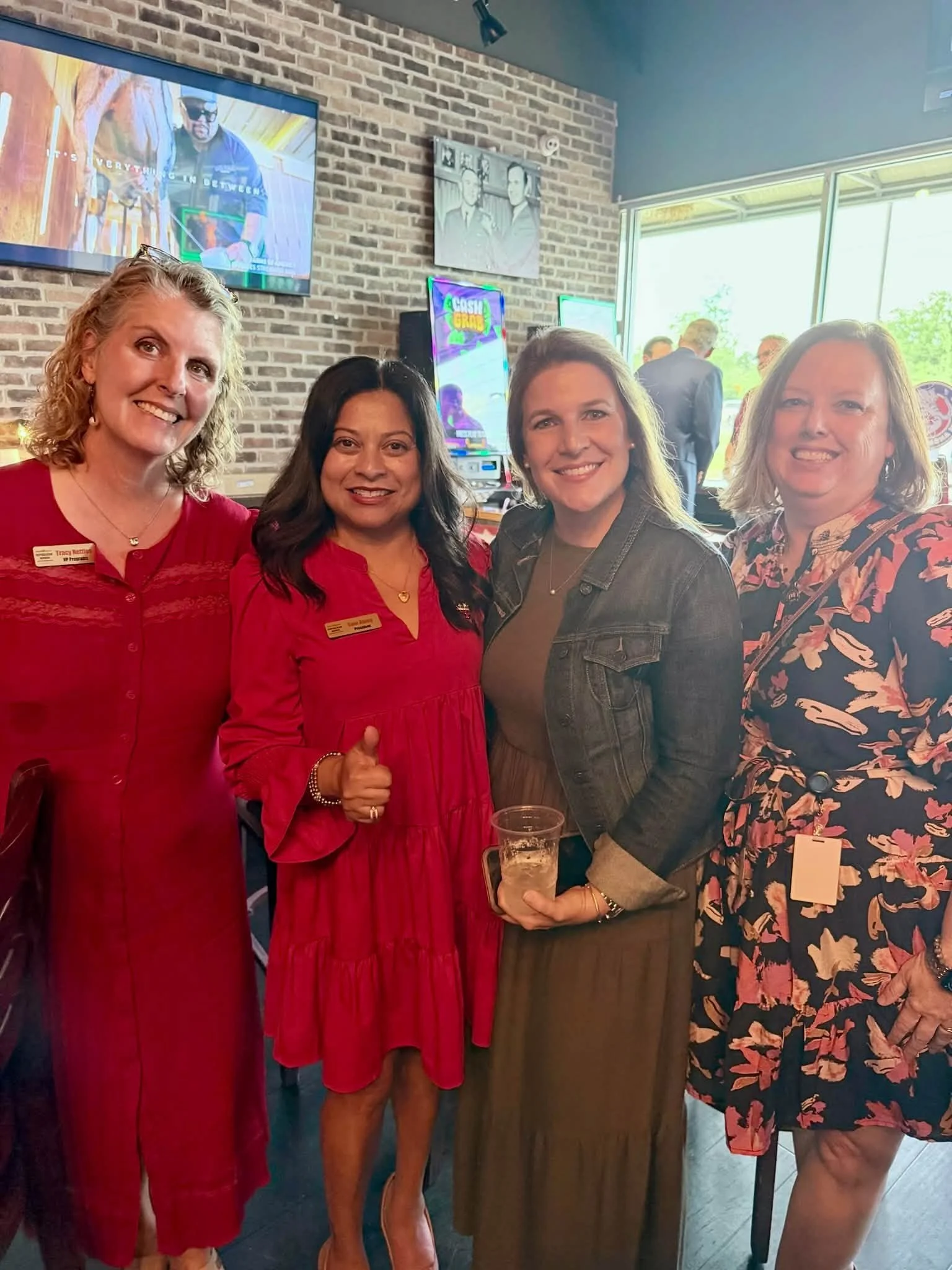 Four women smiling and standing together at an indoor event, with two wearing red dresses and one in a floral dress. One woman is holding a drink. Background includes a brick wall, television screens, and a window with trees outside.