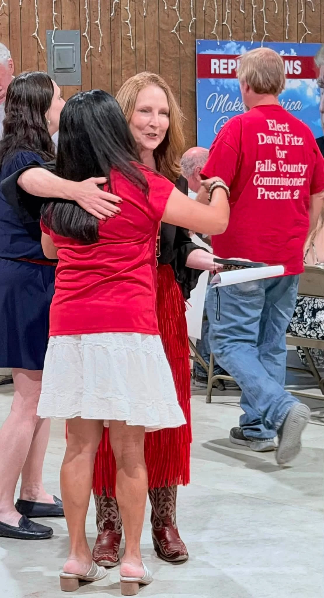 People socializing at an indoor event, with one woman wearing a white skirt and others in casual attire, including a red shirt supporting a political campaign.