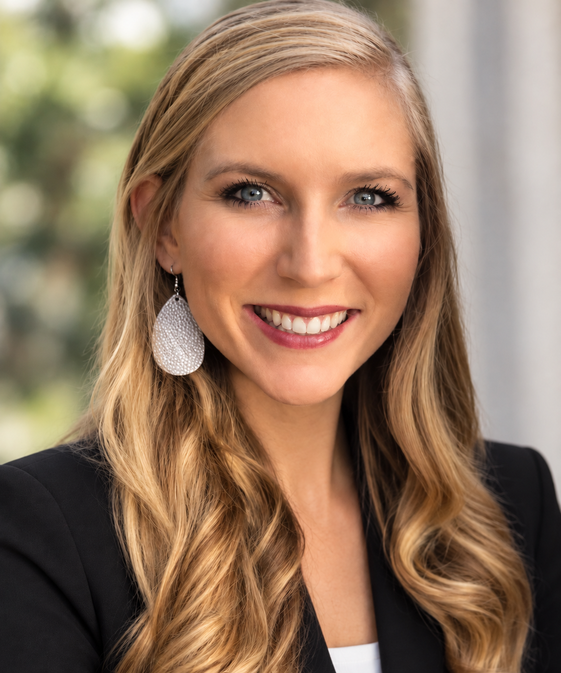 Close-up of a woman with long blonde hair and blue eyes, smiling, wearing a black blazer and large silver earring, standing outdoors with blurred greenery in the background.