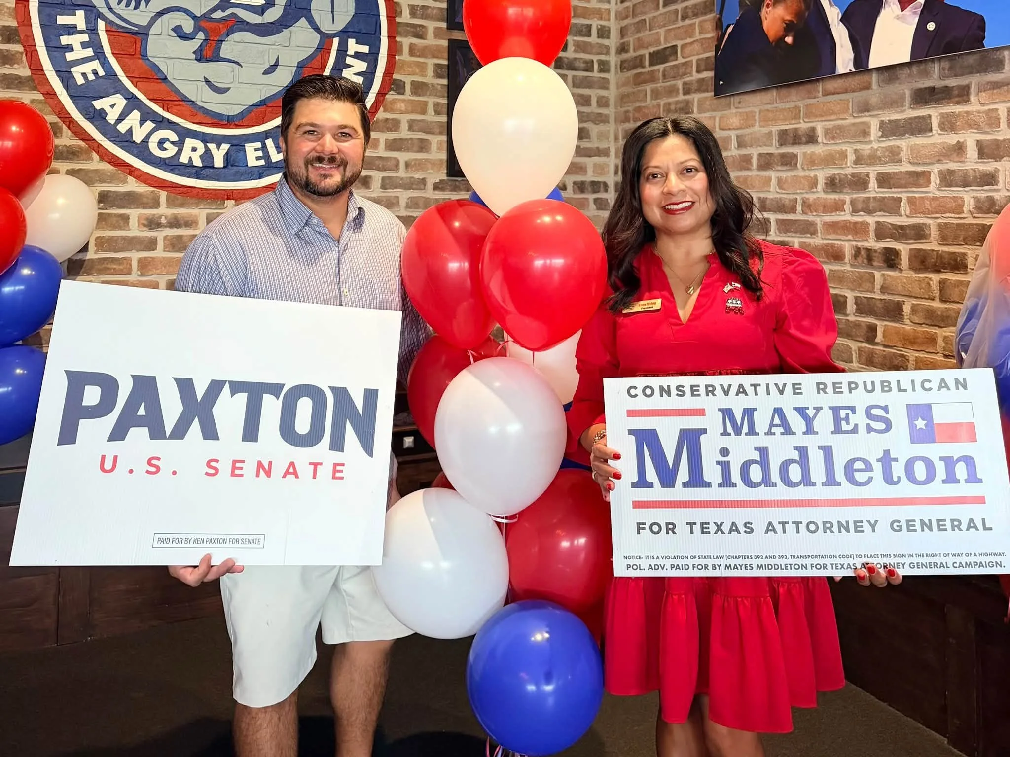 Two people standing in front of a brick wall with balloons and political campaign signs. The man on the left is wearing a light-colored, checkered shirt and holding a sign that reads 'Paid for by Ken Paxton for Senate.' The woman on the right is wear