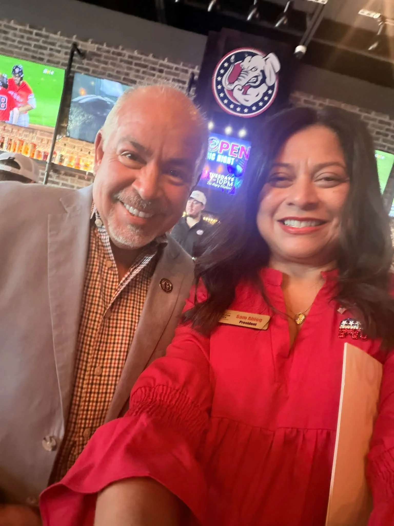 Two smiling people taking a selfie in a bar or restaurant, with television screens and flags in the background.