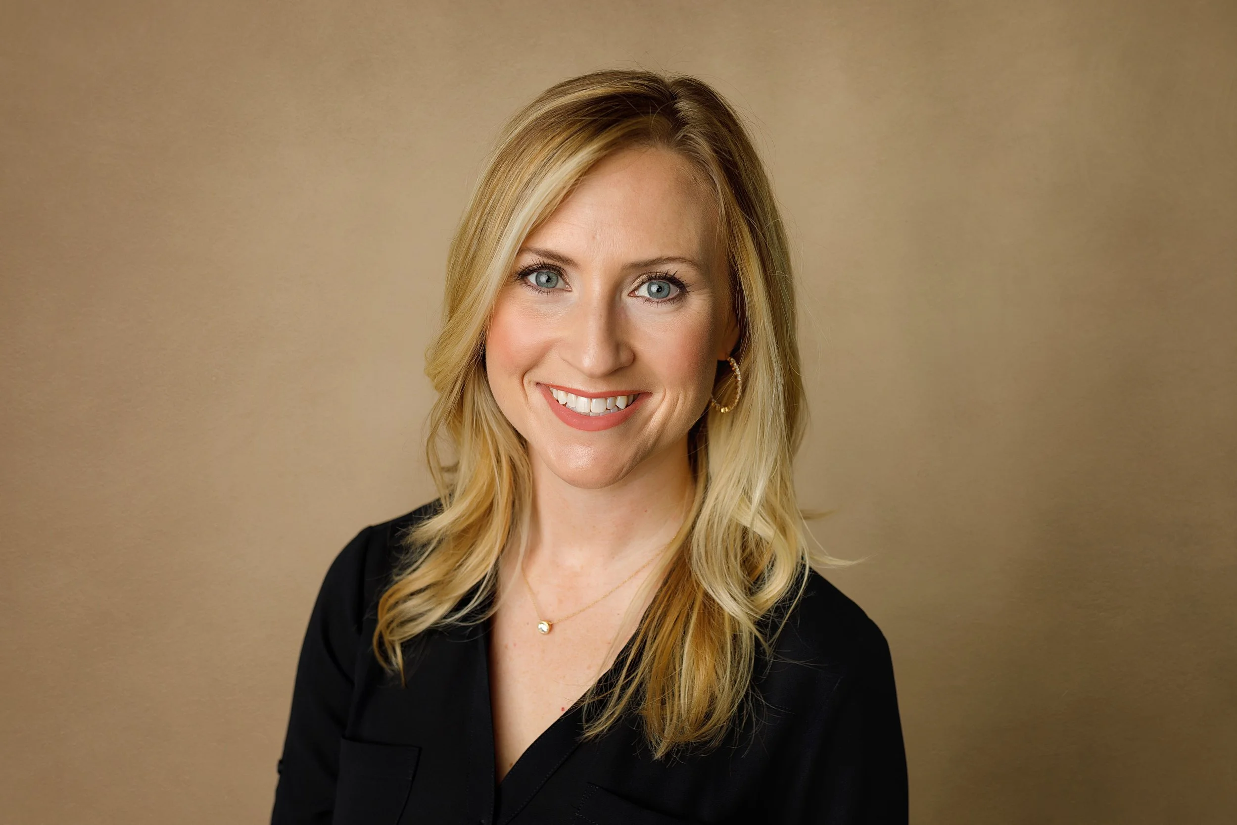 A smiling woman with long blonde hair, blue eyes, wearing a black top, gold hoop earrings, a delicate gold necklace, and makeup, standing against a plain beige background.