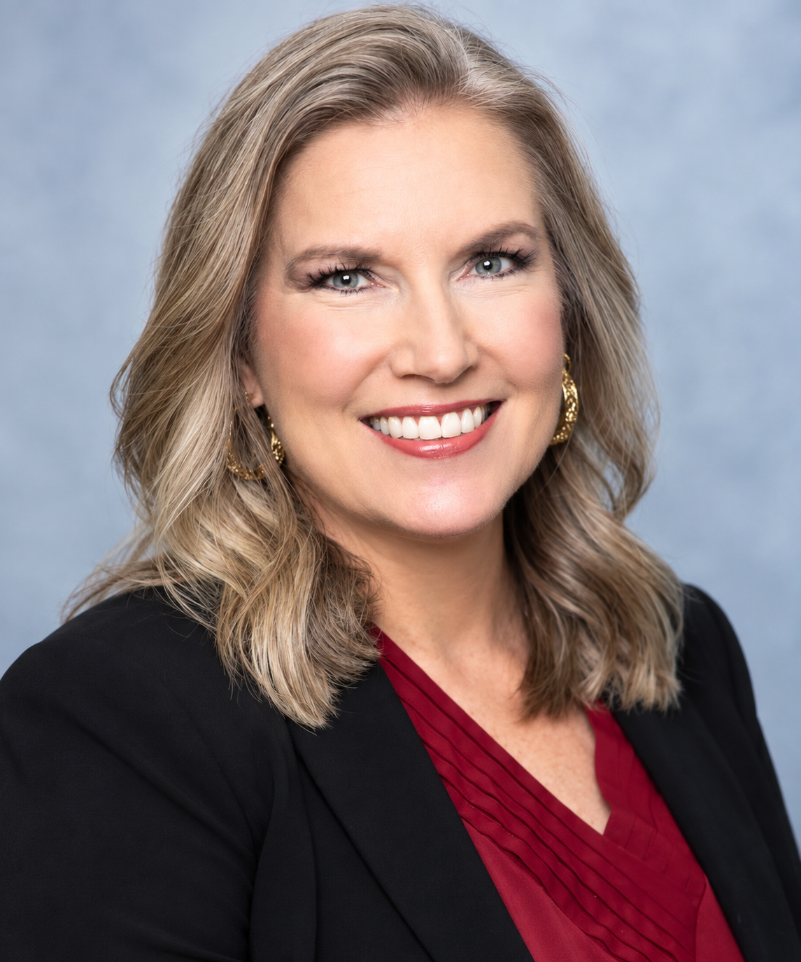 Portrait of a smiling woman with blonde, shoulder-length wavy hair, wearing a black blazer over a red blouse with pleats, gold earrings, and natural makeup, against a light blue background.