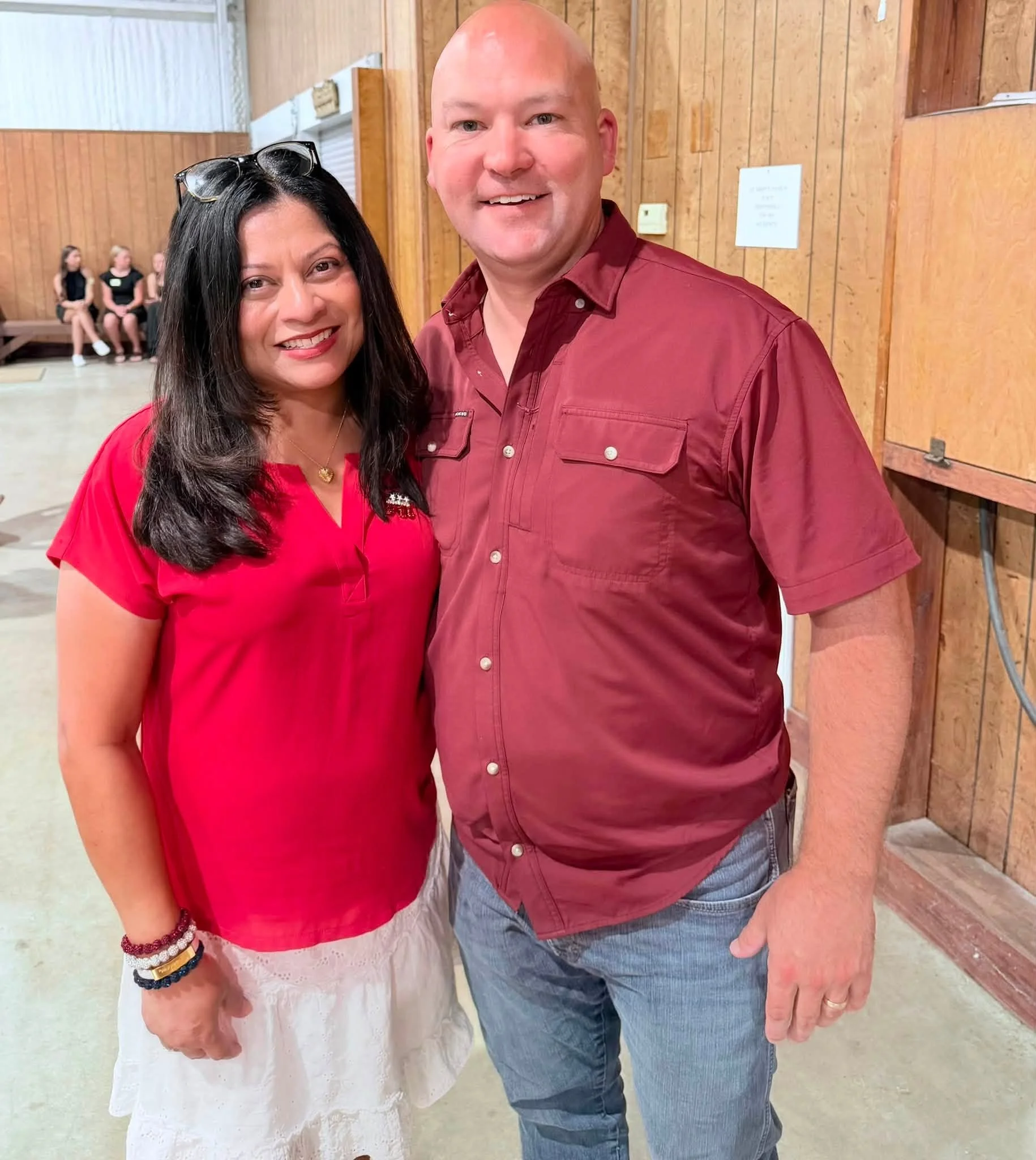 A woman and a man standing together indoors, smiling at the camera. The woman has long dark hair, wears a red top, white lace skirt, and accessories. The man is bald, wearing a red button-up shirt and blue jeans. In the background, there are wooden w