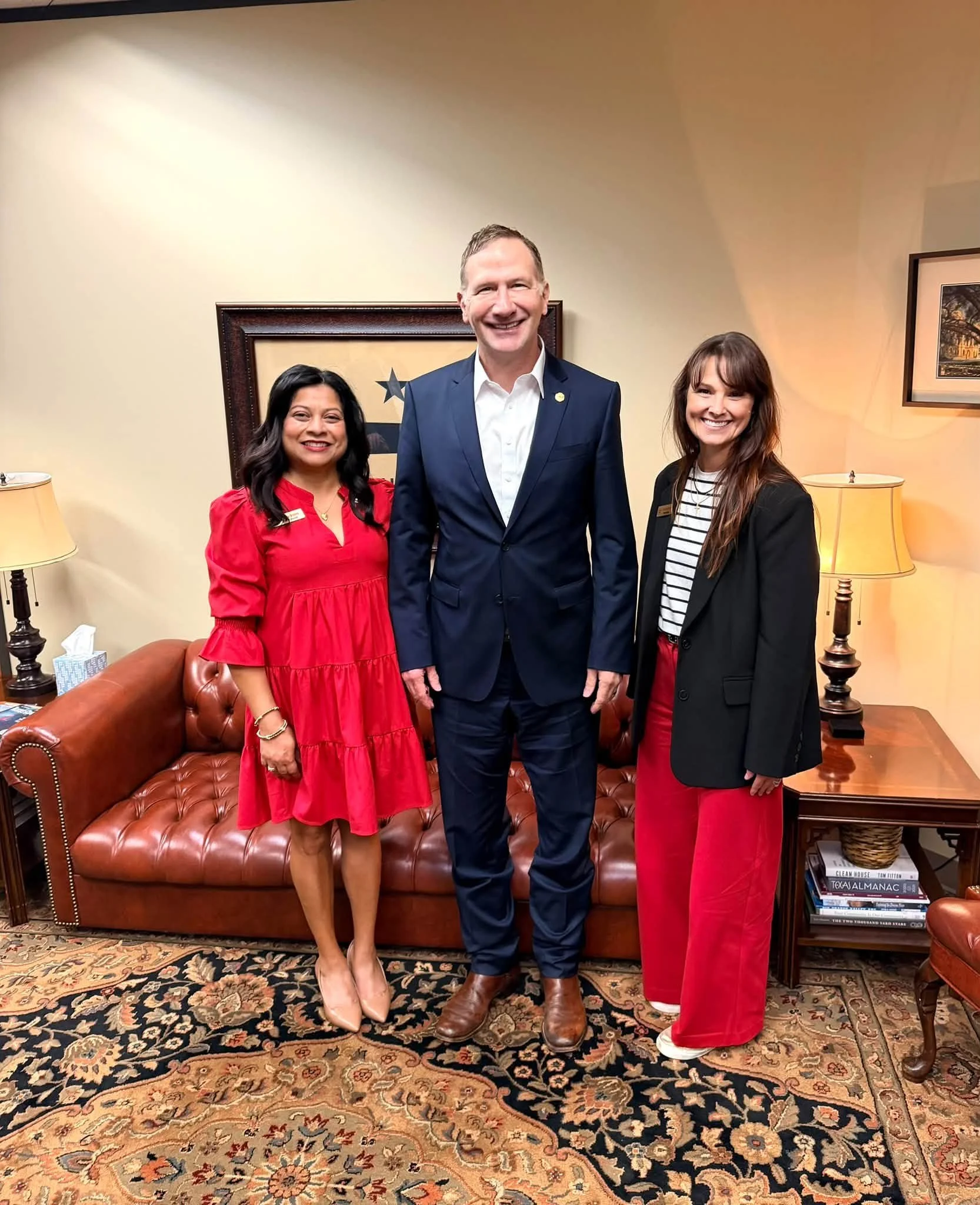 Three people, two women and a man, standing together in an office. The woman on the left wears a red dress and beige shoes. The man in the middle wears a navy suit and brown shoes. The woman on the right wears a black blazer, striped shirt, red pants