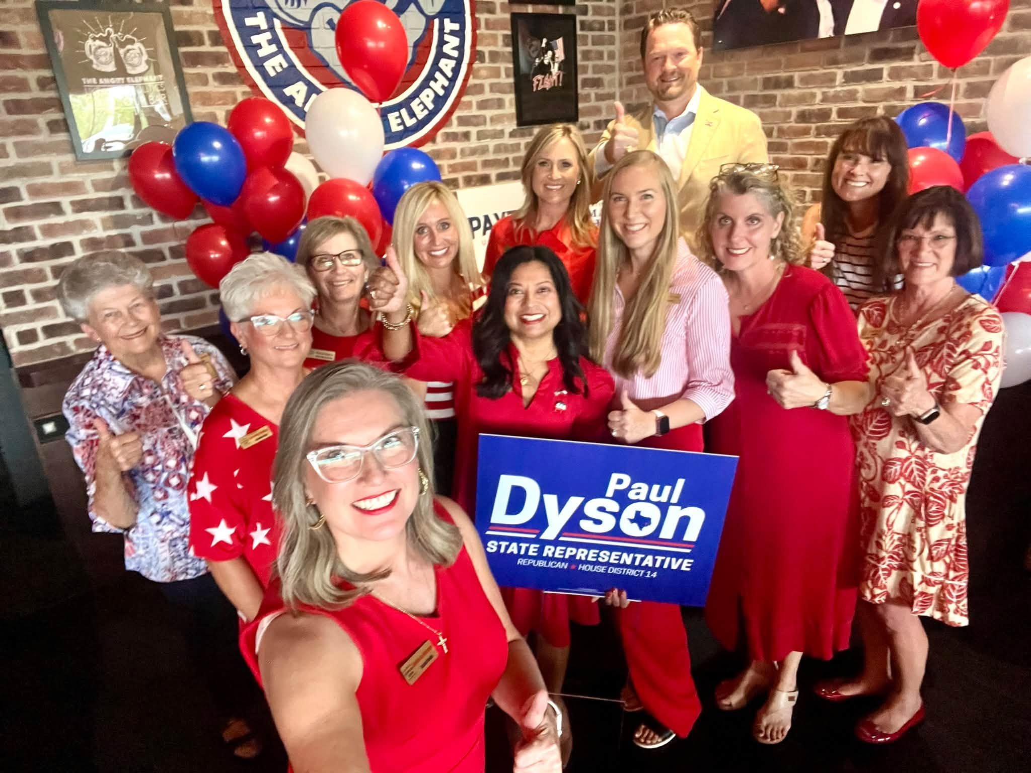 Group of people celebrating, holding a sign supporting Paul Dyson for State Representative. The group is dressed in red, white, and blue, with balloons in similar colors. They are smiling and giving thumbs-up in front of a brick wall with political b