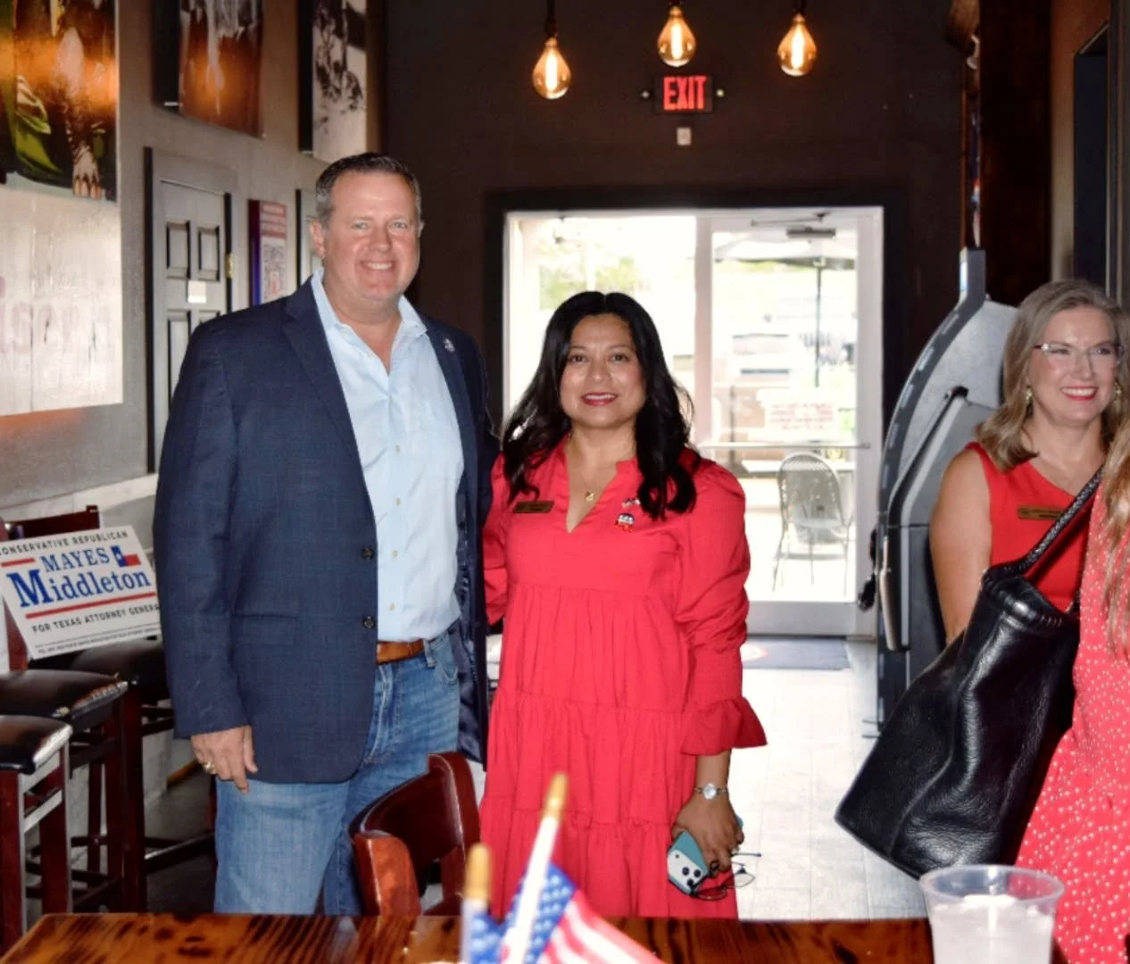 A group of three people standing inside a restaurant. A man and a woman in the center are smiling; the woman is wearing a red dress, and the man is in a blue suit jacket and light blue shirt. On the right, a woman with glasses and blonde hair, wearin
