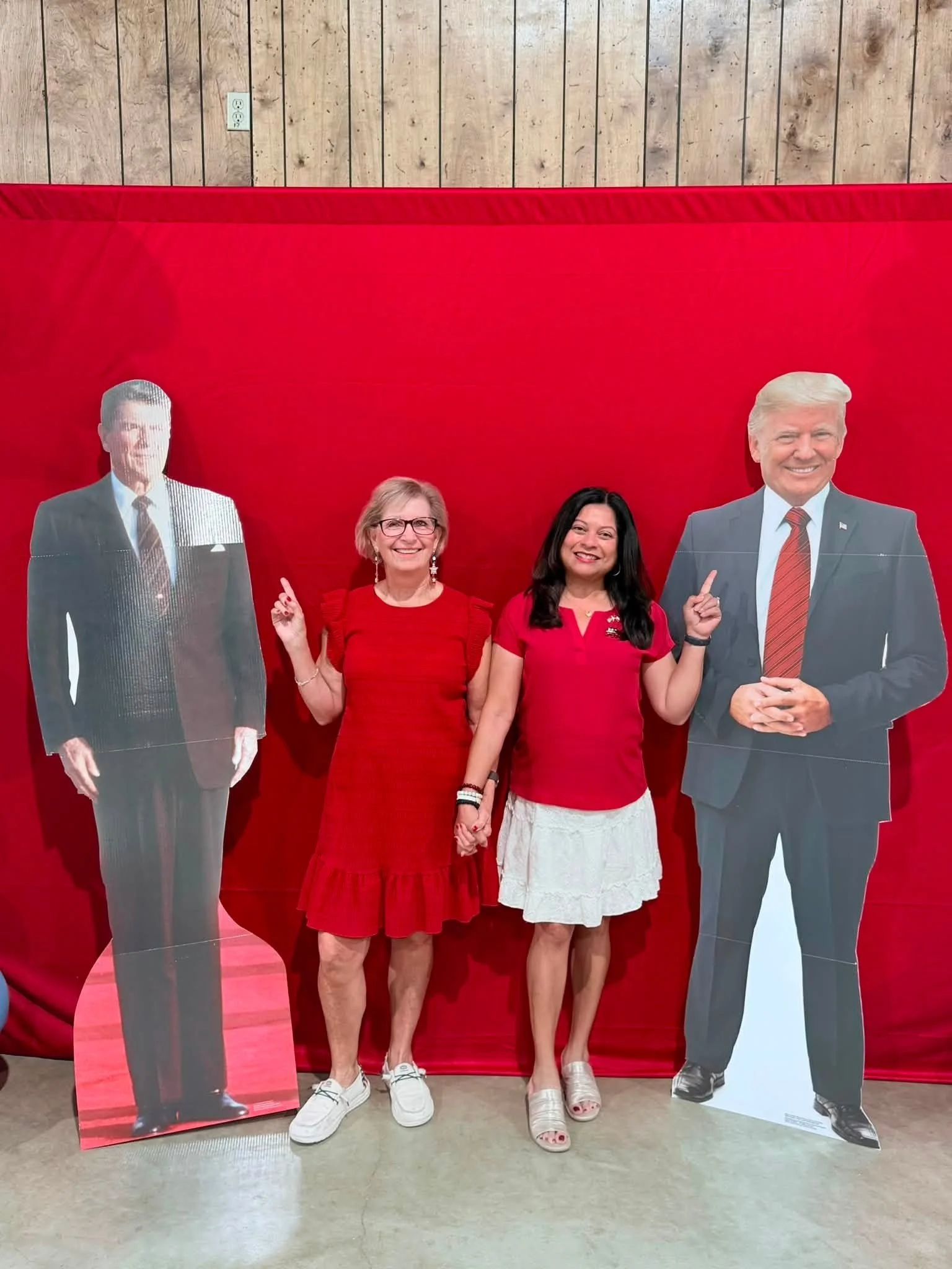 Two women stand hand in hand in front of life-sized cardboard cutouts of former President Donald Trump in a room with wooden walls and a red backdrop.