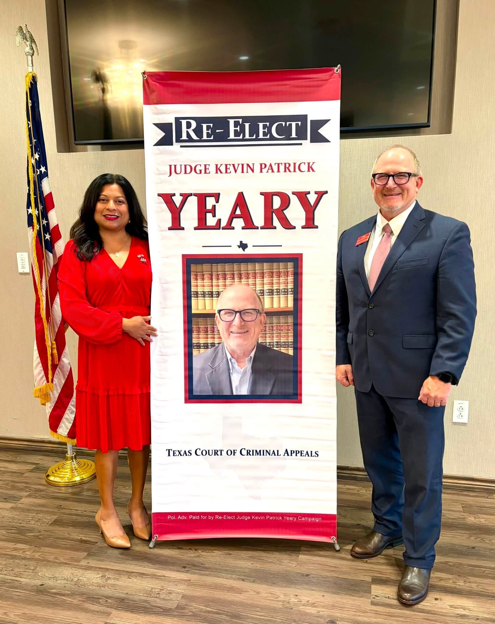 Two people standing next to a Re-Elect Judge Kevin Patrick Yeary campaign banner, with an American flag to the left. The woman is wearing a red dress and beige heels, and the man is dressed in a blue suit with a pink tie and brown shoes. They are in 