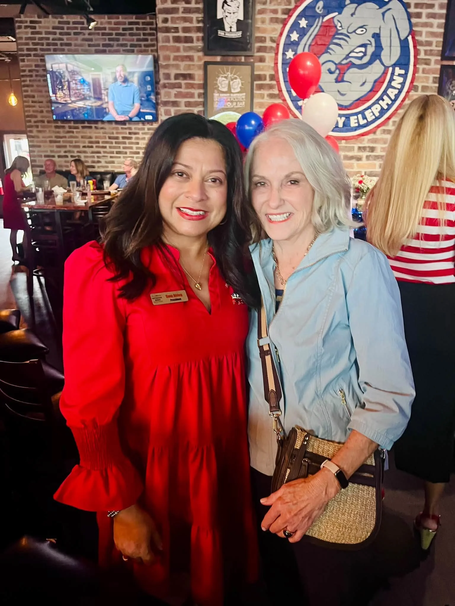 Two women standing together in a restaurant, one with dark hair wearing a red dress and name tag, and the other with white hair wearing a light blue jacket. There are red, white, and blue balloons and a large sports team logo on the brick wall behind