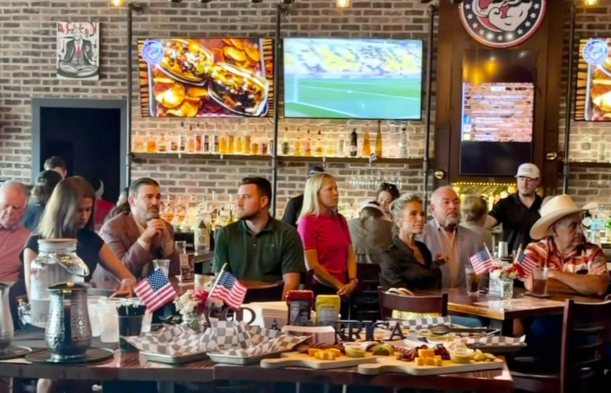 People dining at a restaurant with tomato and cheese platter on table, brick wall with TV screens, patriotic decorations, and American flags.