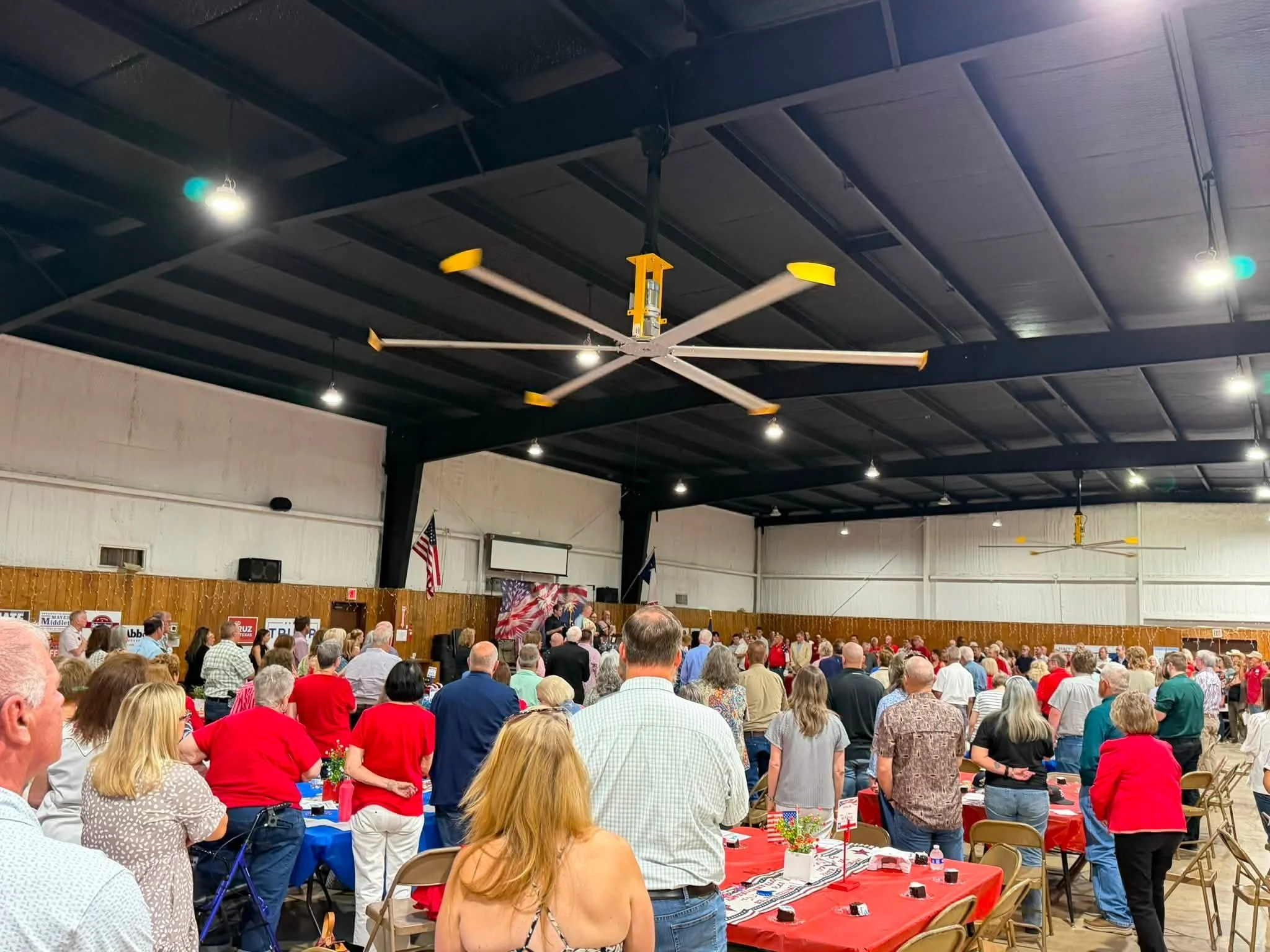 A large crowd of people gathered inside a spacious room with a high ceiling and dark beams, attending a political event or rally. There are tables covered with red, white, and blue tablecloths, decorated with small country flags and flowers. Some att