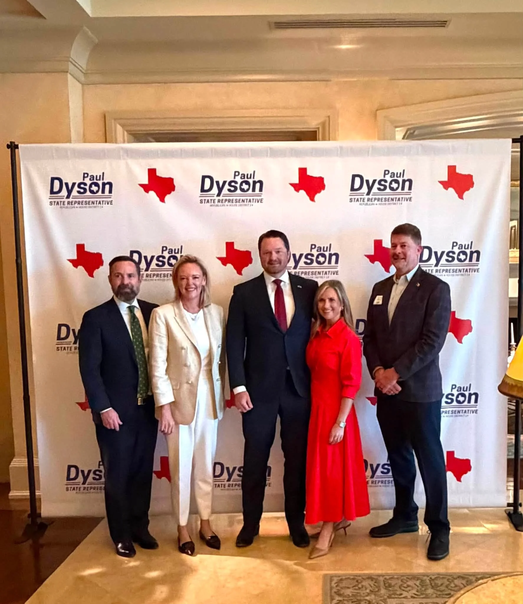 Group of six professionally dressed people standing in front of a campaign backdrop with the text "Paul Dyson State Representative" and a red Texas shape logo, in an indoor setting.