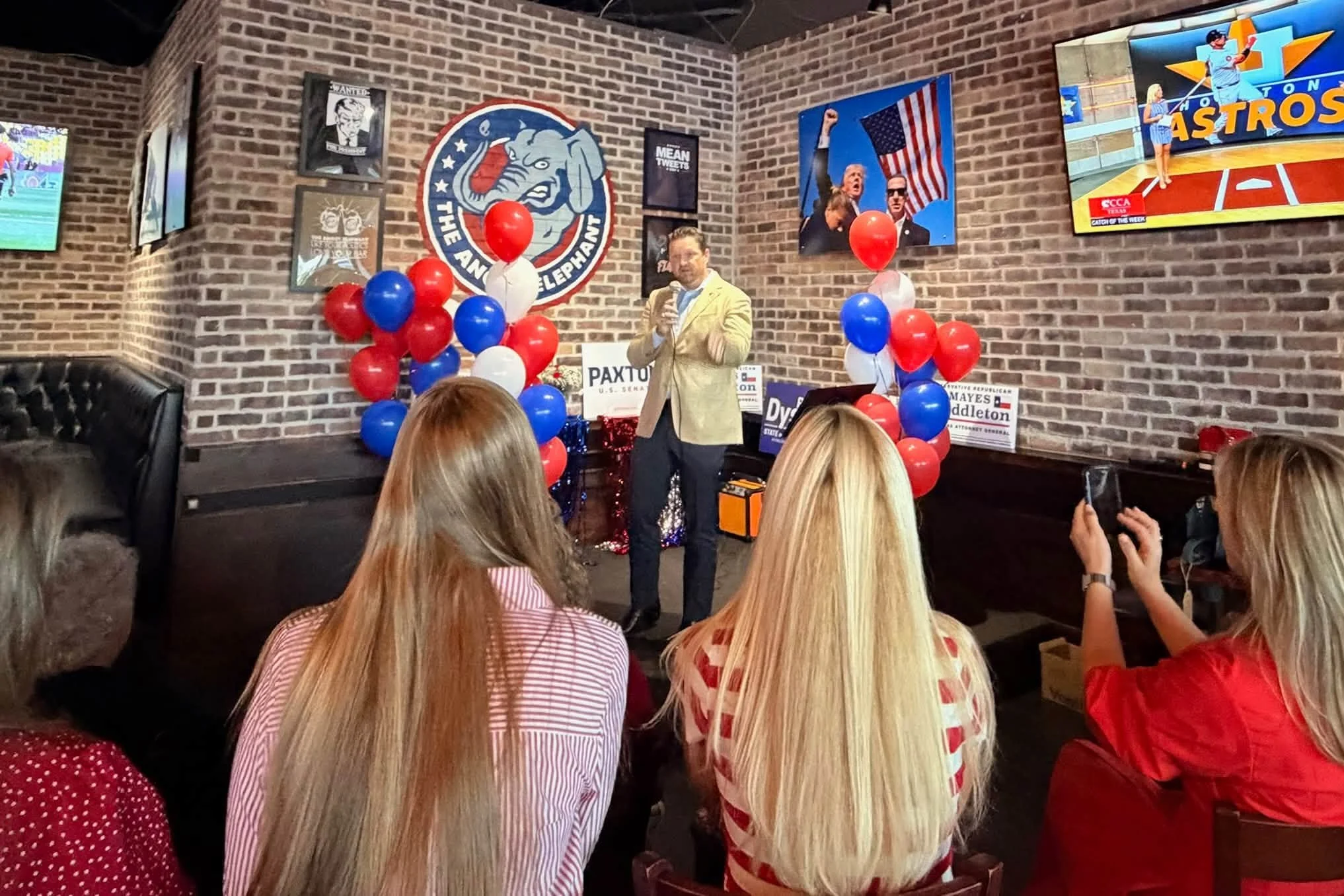 A man in a yellow jacket speaking at an event with red, white, and blue balloons in the background. Several people are seated facing him, and there are patriotic and sports-themed posters and a flat-screen TV on the brick wall behind him.