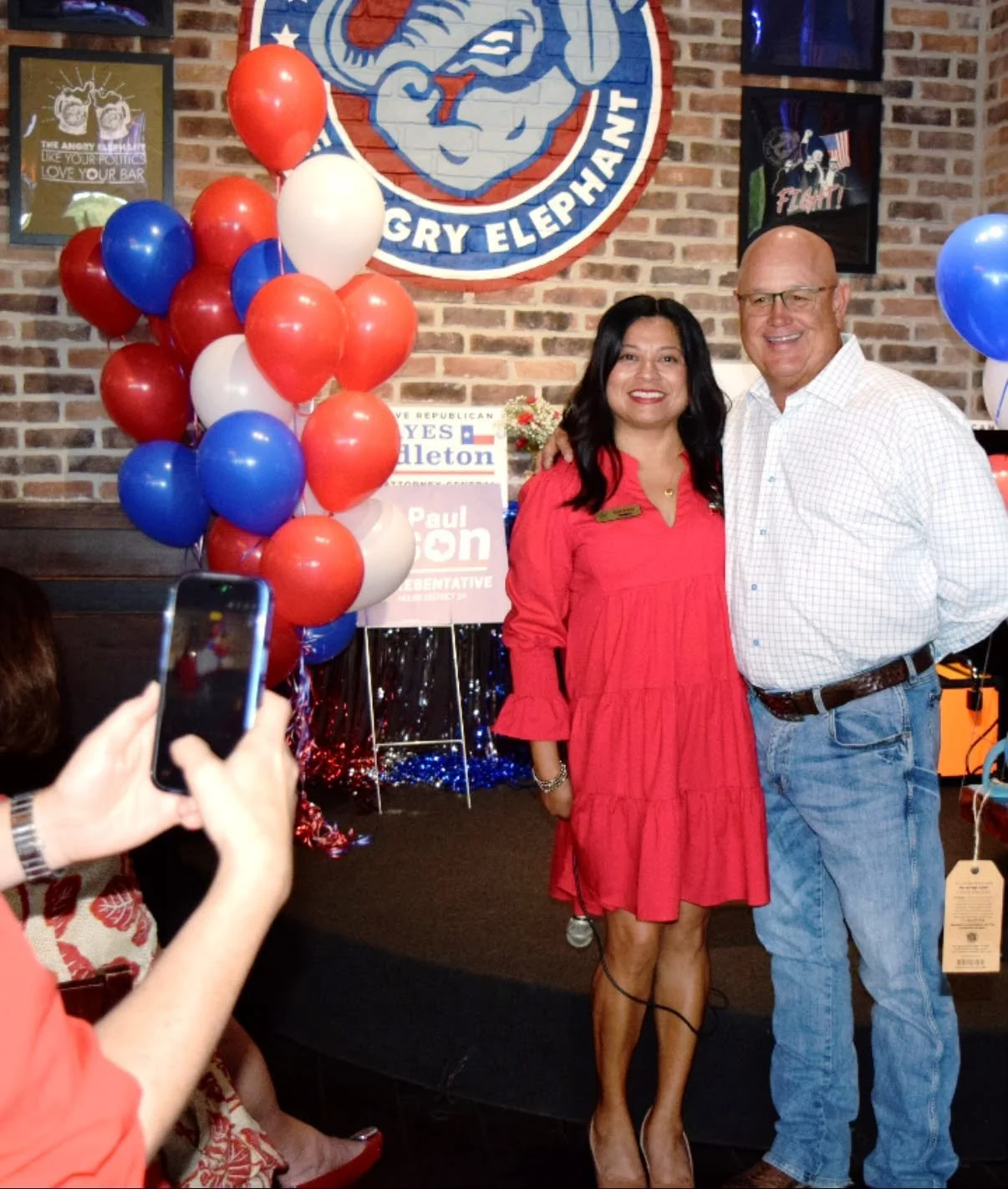 Two people standing and posing for a photo at a political event. The woman is wearing a red dress, and the man is wearing a white checkered shirt and jeans. Behind them, there is a brick wall with political posters, balloons in red, white, and blue, 