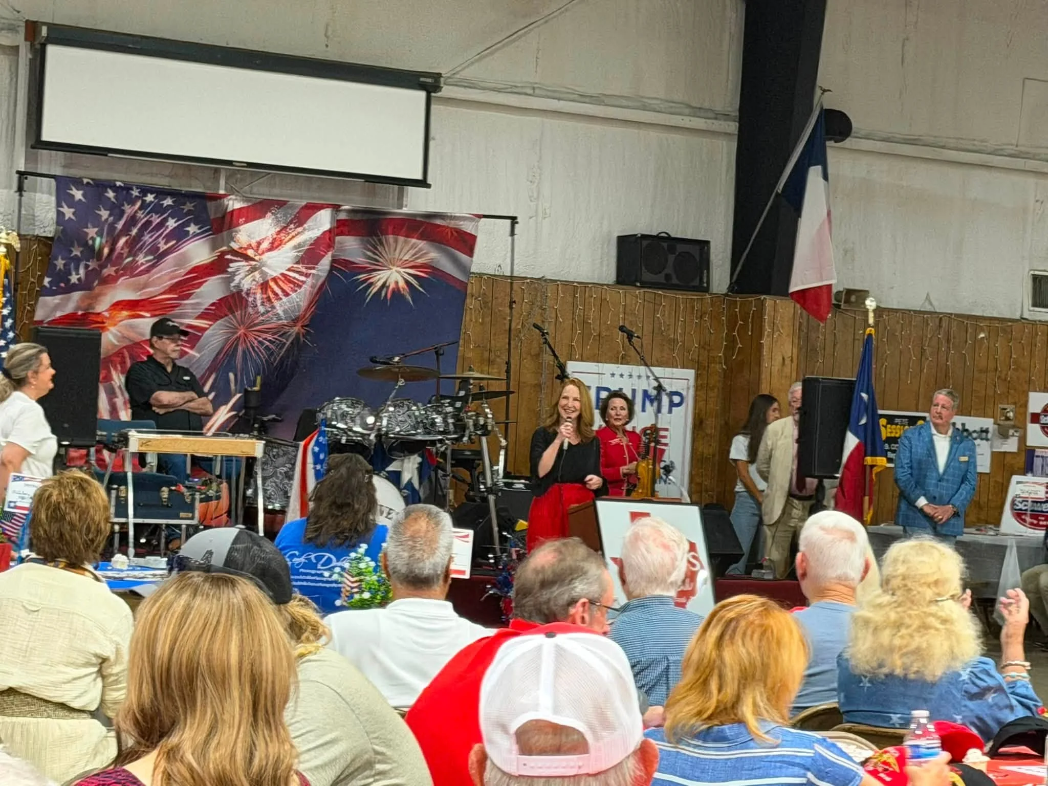 A crowd of people gathered at an indoor event with banners and decorations, including American flag-themed banners and a 'TRUMP' sign. There is a stage with musicians and speakers, and the audience is seated at tables.
