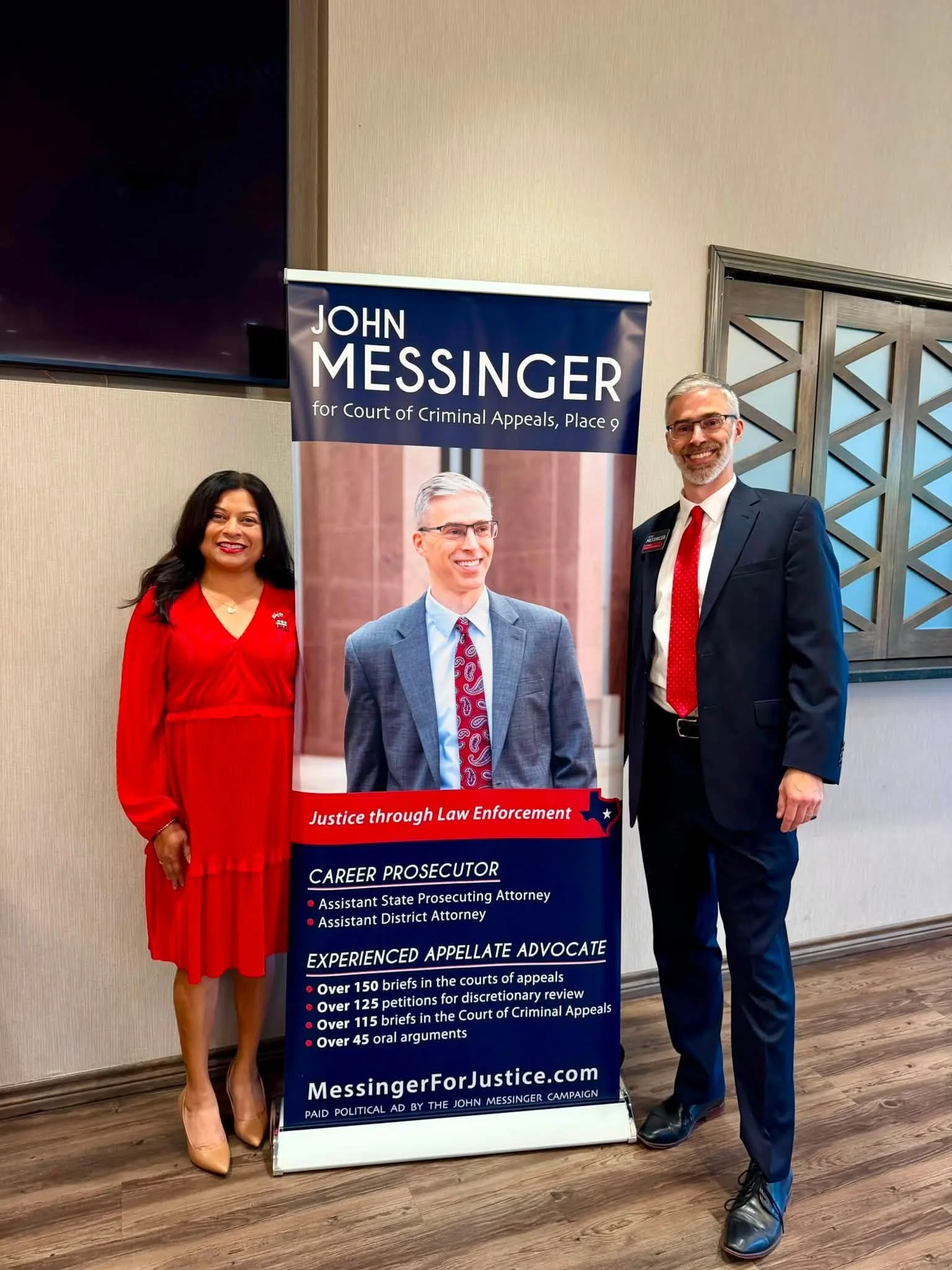 Two people standing next to a campaign poster for John Messinger, a candidate for Court of Criminal Appeals. The woman on the left is wearing a red dress and beige heels, and the man on the right is in a dark suit with a red tie and black shoes, both