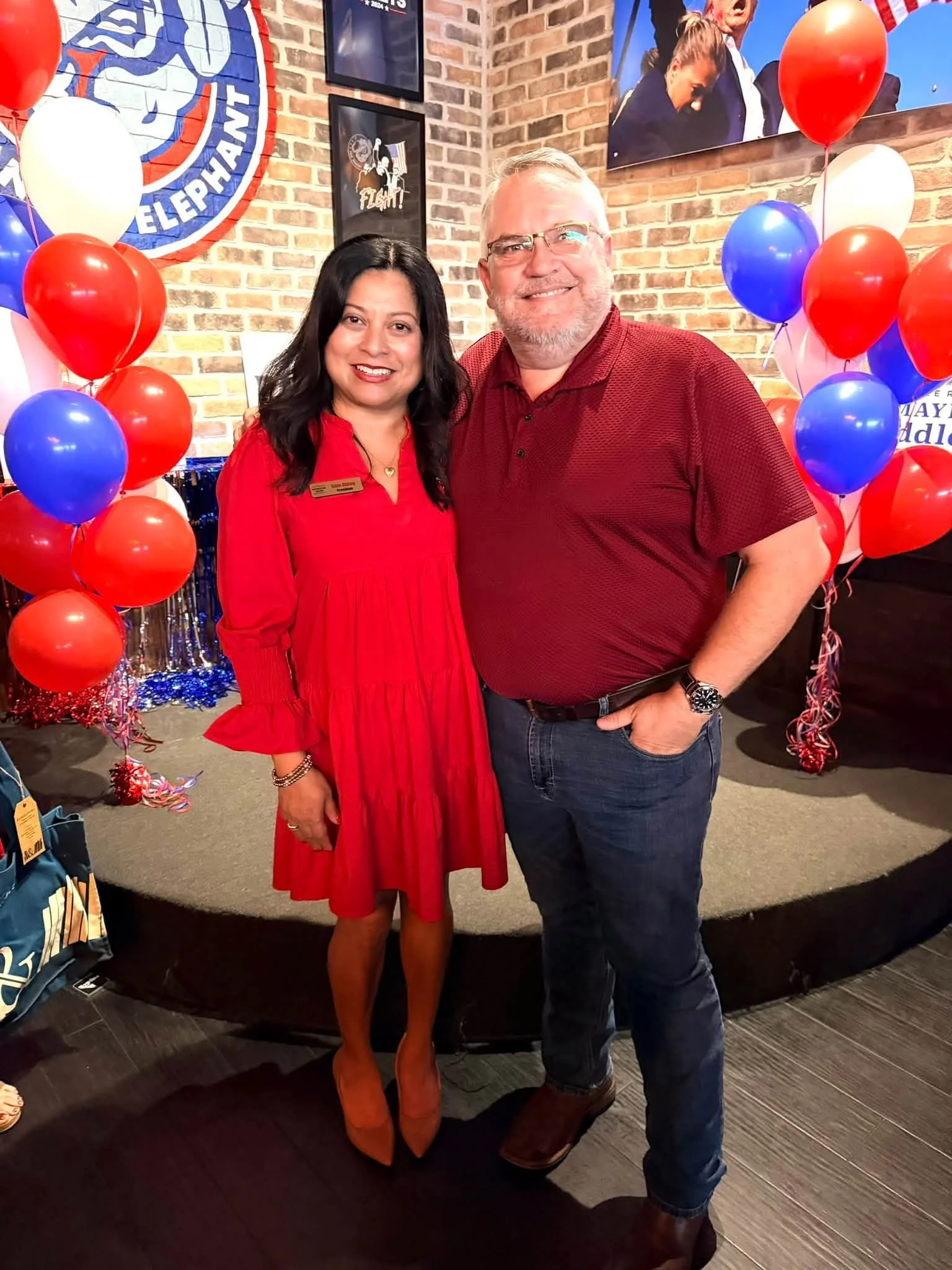 A woman and a man standing together at a celebration. The woman is wearing a red dress and tan boots, and the man is wearing a red shirt and jeans. There are red, white, and blue balloons and decorations around them. The background features a brick w