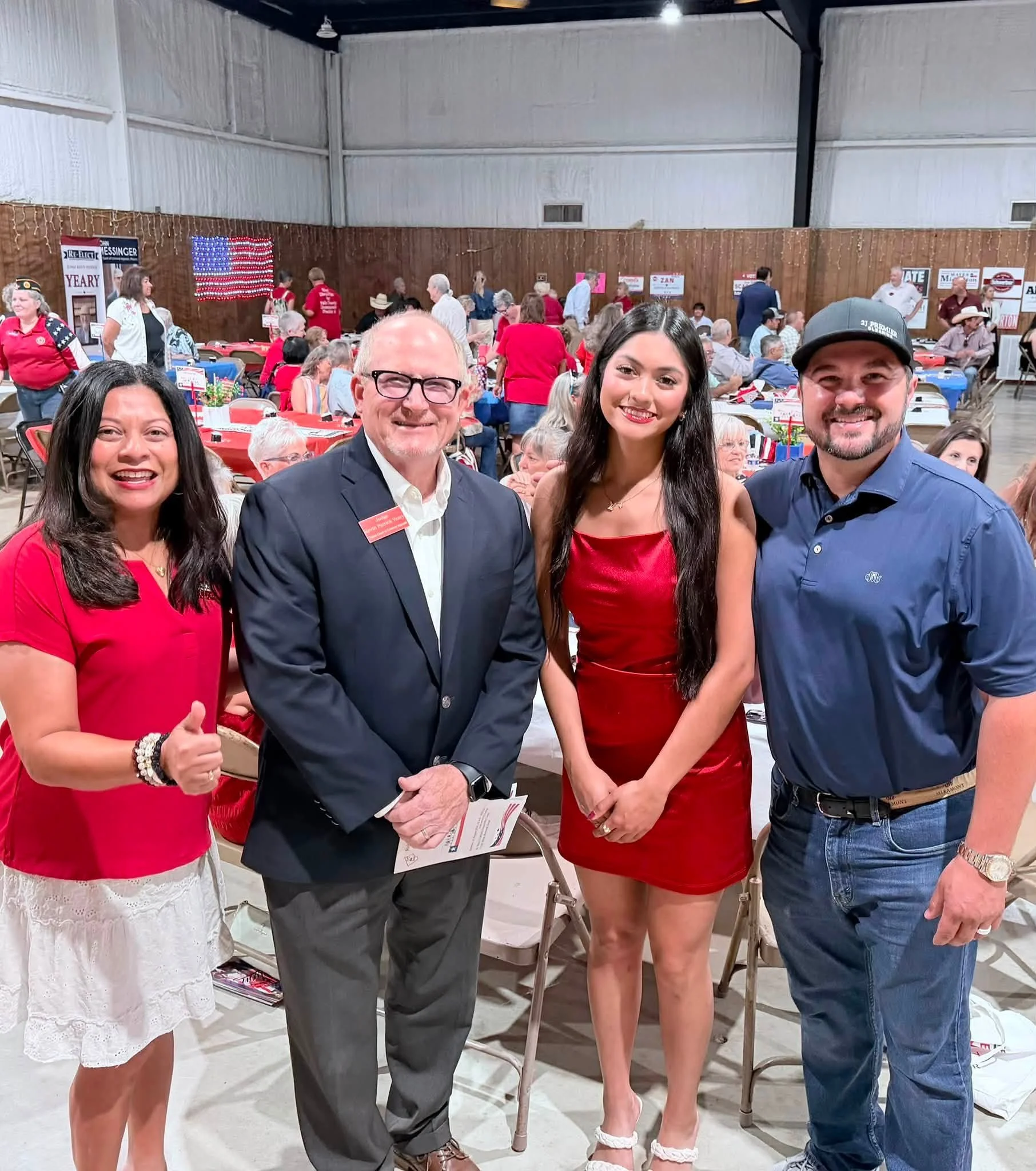 A group of five people posing for a photo at a crowded indoor event with tables and banners in the background. They are smiling and dressed in semi-formal attire, some wearing red, white, and blue, indicating a patriotic theme.