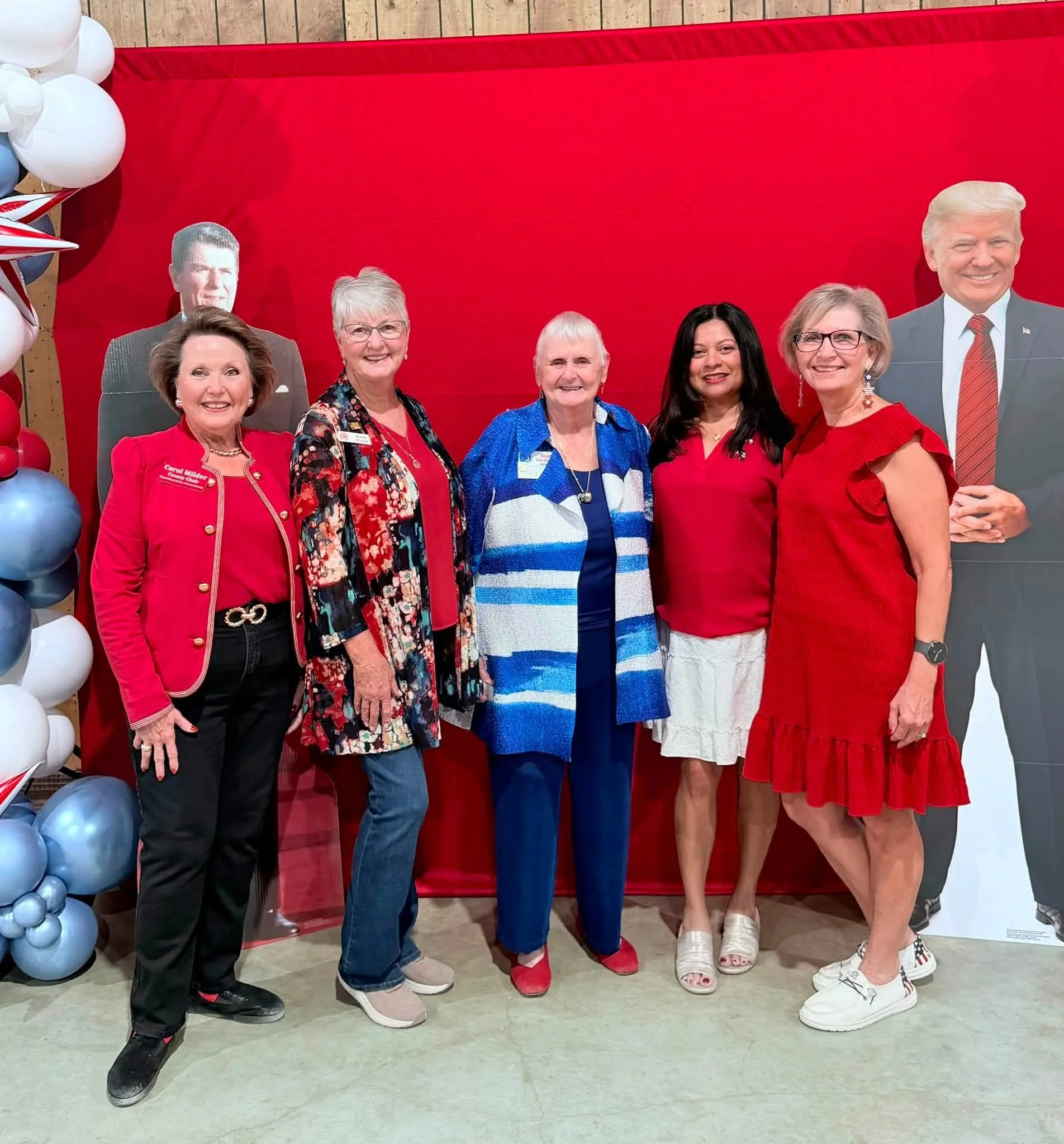 Group of six women and one man standing in front of a red backdrop with balloon decorations on the side. The women are dressed in red and colorful outfits, and the man, who is positioned at the far right, is wearing a suit and tie. They are smiling f