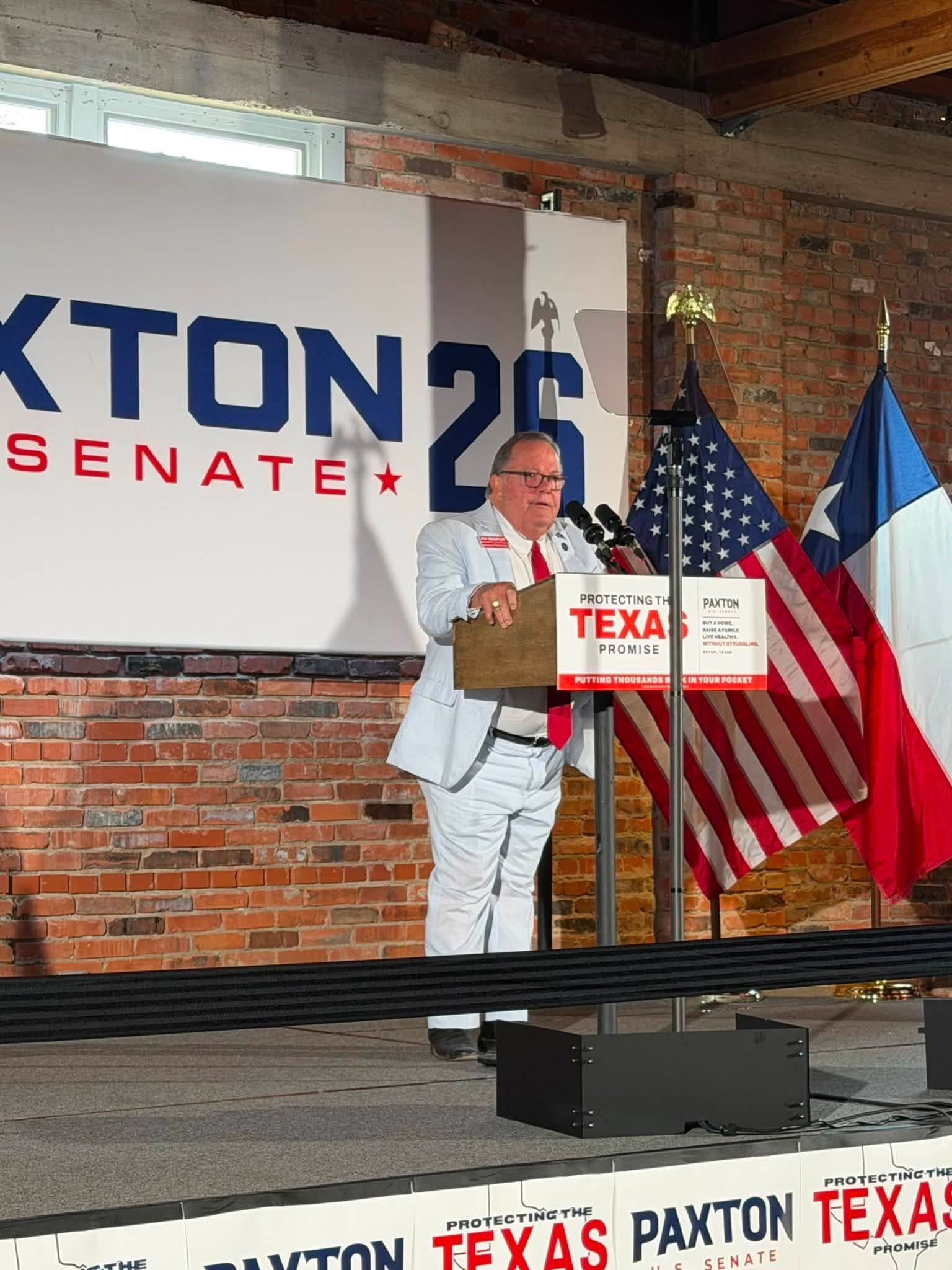 A man dressed in a white suit with a red tie and glasses stands at a podium with microphones, giving a speech. Behind him, there are American flags, a large campaign banner reading 'KTON 26' and 'SENATE,' and a brick wall with windows and wooden beam