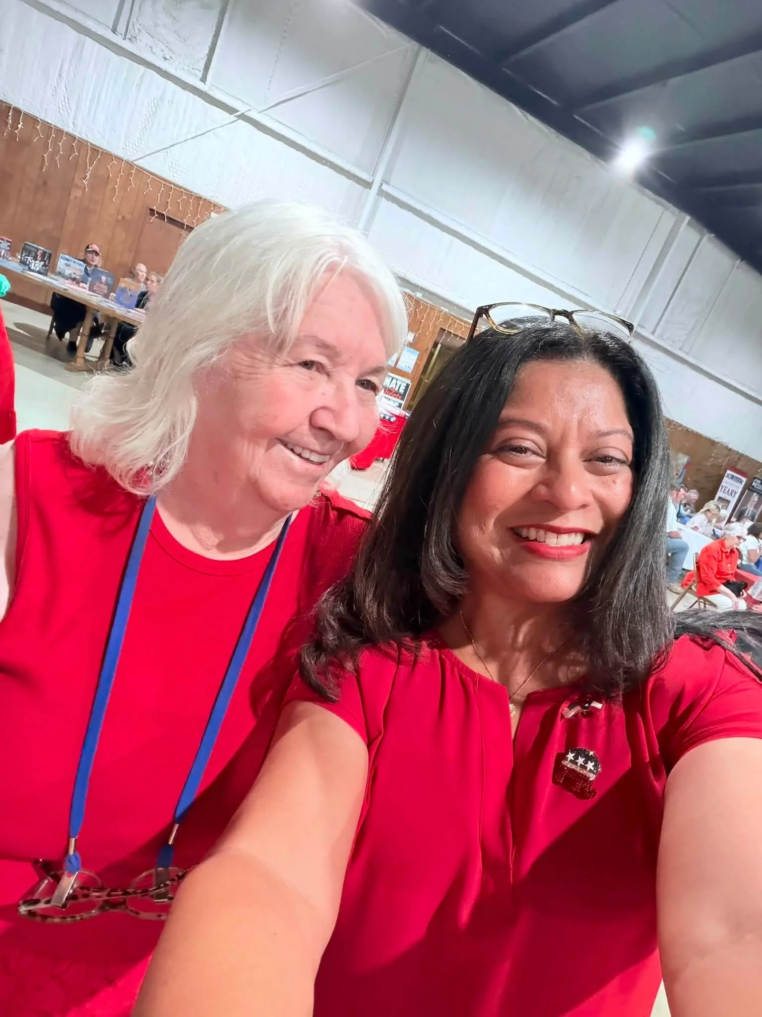 Close-up of two women smiling, wearing red clothing, with a background of seated people at tables in a hall.
