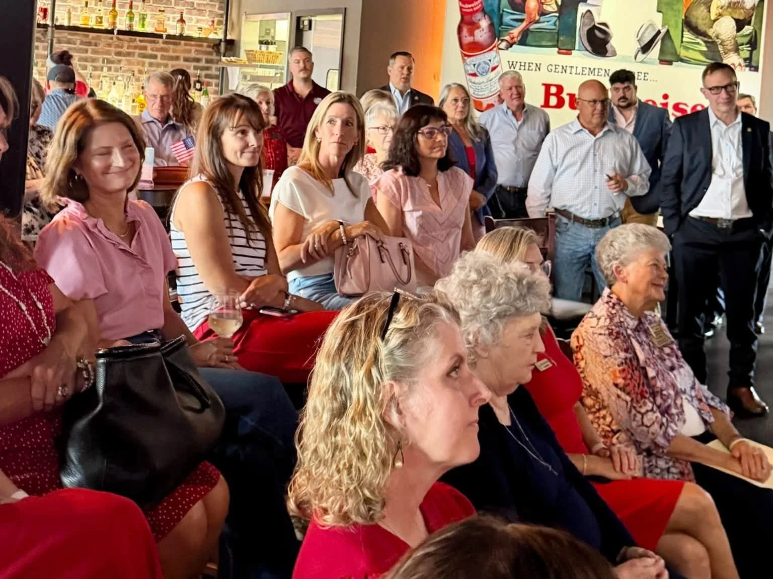 A group of people, mainly women, attending an event in a restaurant or bar. Some are seated, and some are standing, watching something in front of them. The background features a brick wall, bottles on shelves, and a retro advertisement poster.