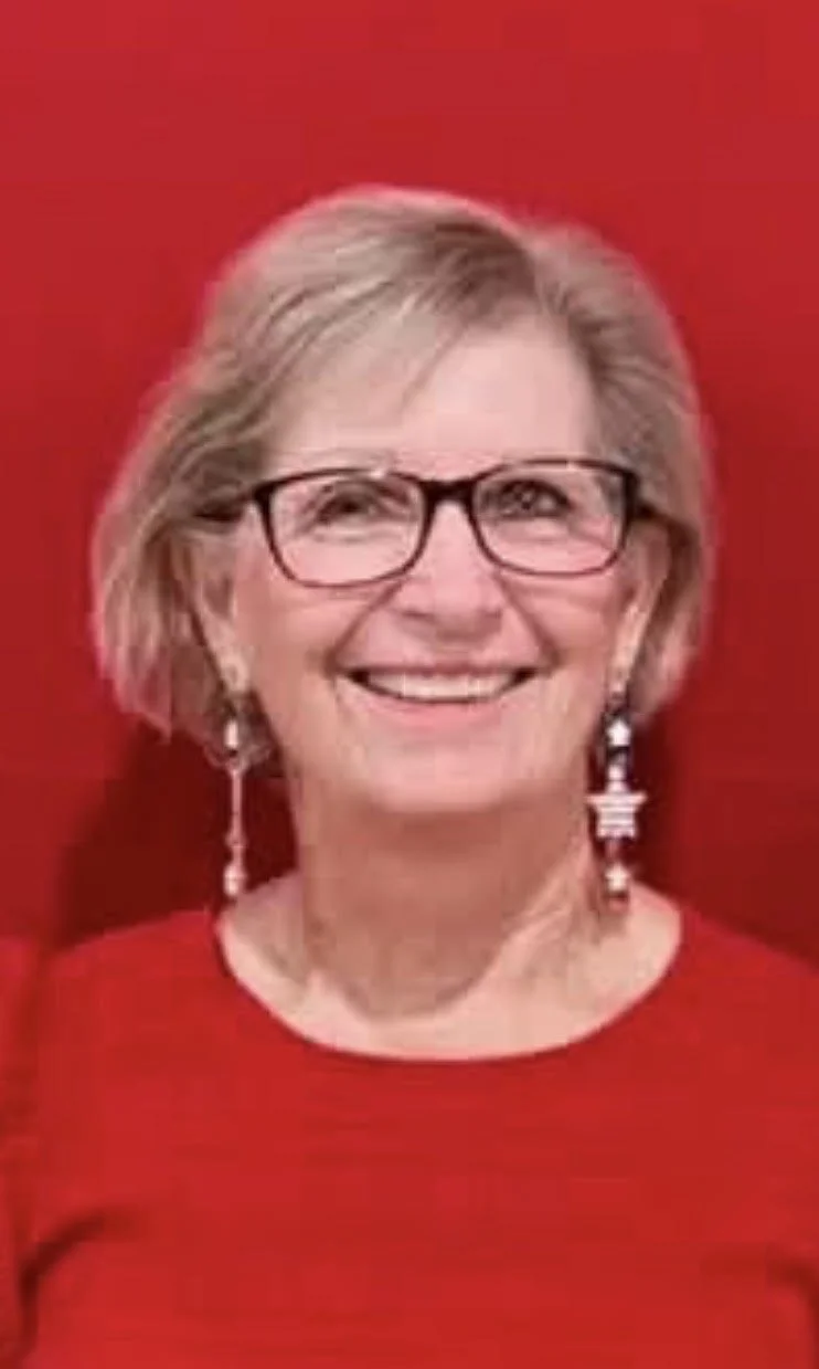 A woman with blonde hair, glasses, and earrings smiling against a red background.