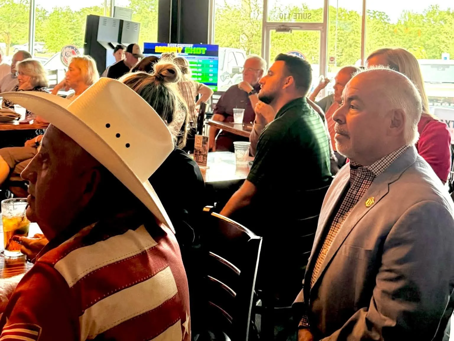 People seated at a bar or restaurant, some engaged in conversation, with a large TV screen displaying a sports betting or casino game behind them, and large windows with greenery outside.