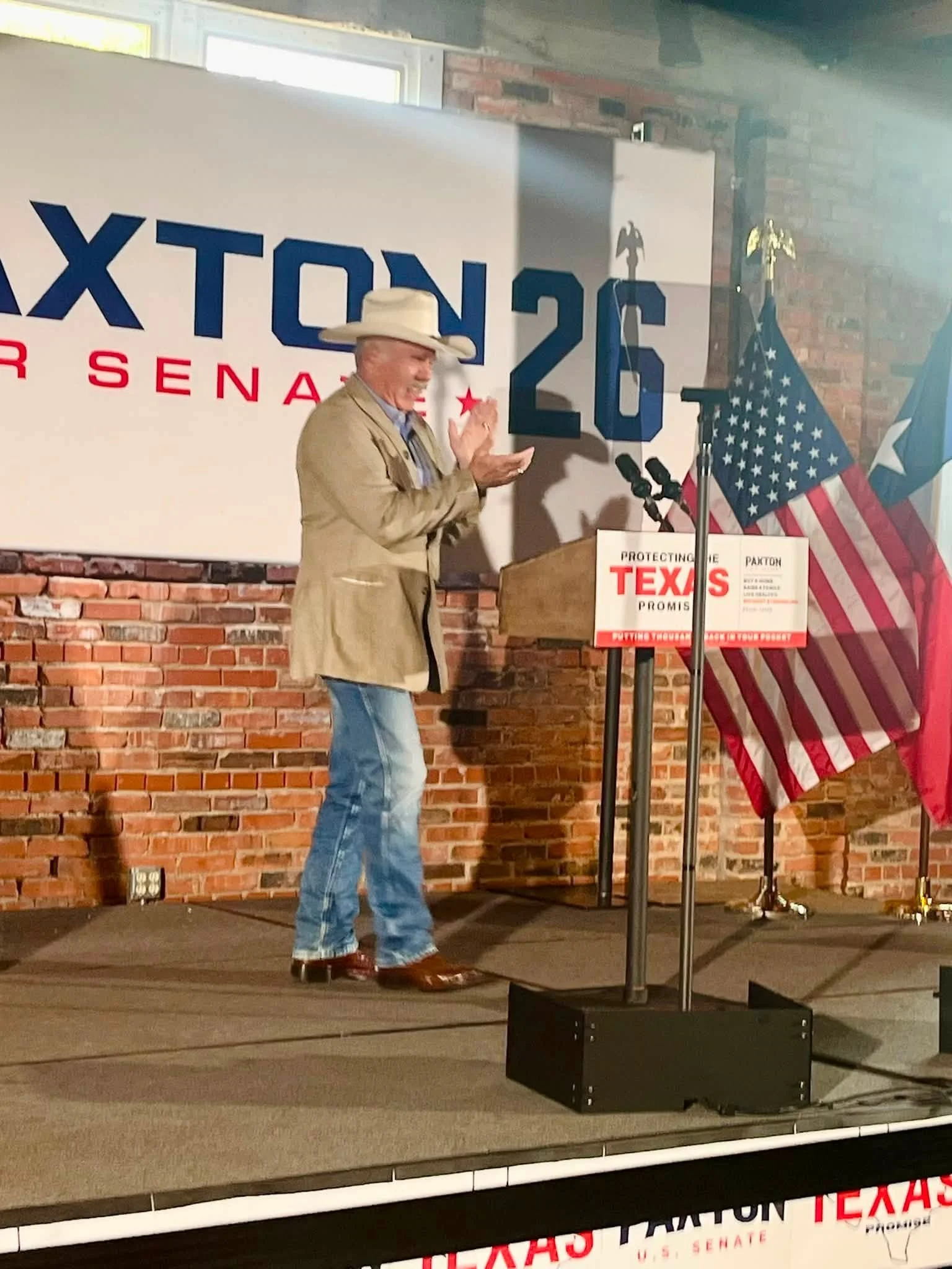 A man in cowboy attire, including a cowboy hat, standing on a stage in front of a campaign sign with the words 'Texan for Senate' and the number 26. He is smiling and gesturing with his right hand. Behind him, there are American and Texas flags.