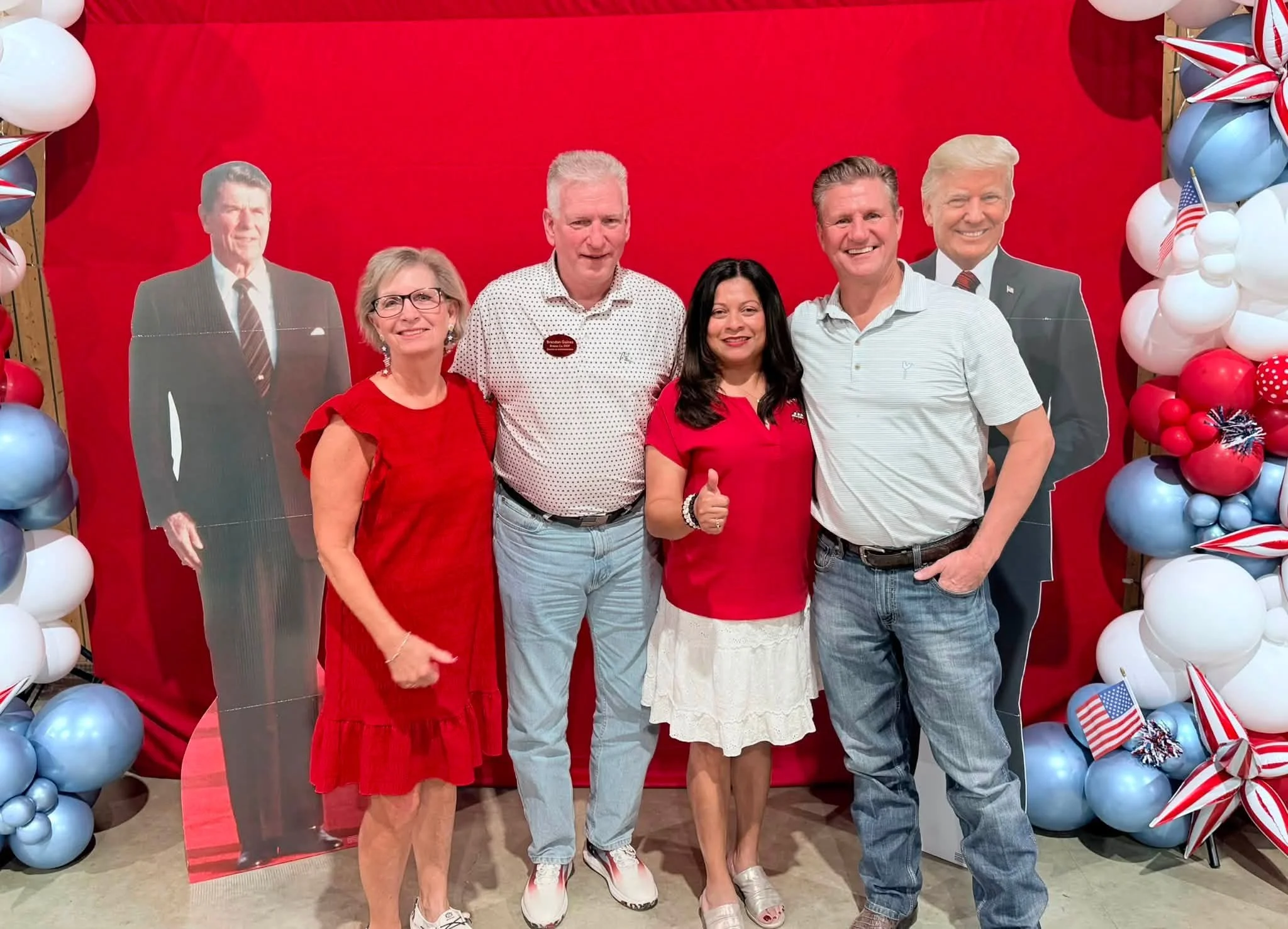 A group of five people posing in front of a patriotic red backdrop with balloons and American flag decorations. They are smiling, and some are giving a thumbs-up.