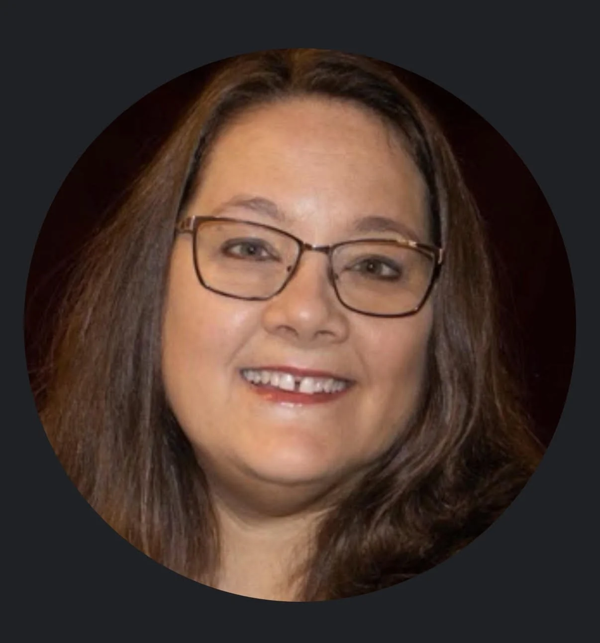 Close-up of a woman with shoulder-length brown hair, wearing glasses, smiling, with a dark background.