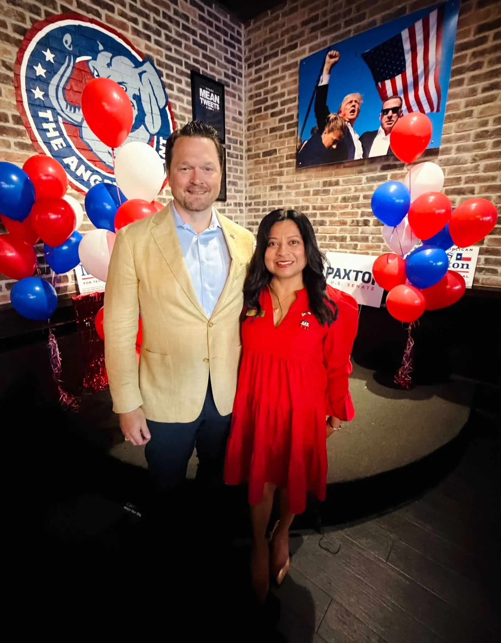 A man and woman standing in front of a brick wall decorated with red, white, and blue balloons, political posters, and a large photograph of former U.S. President Donald Trump at an event supporting U.S. Senate candidate Christophe.