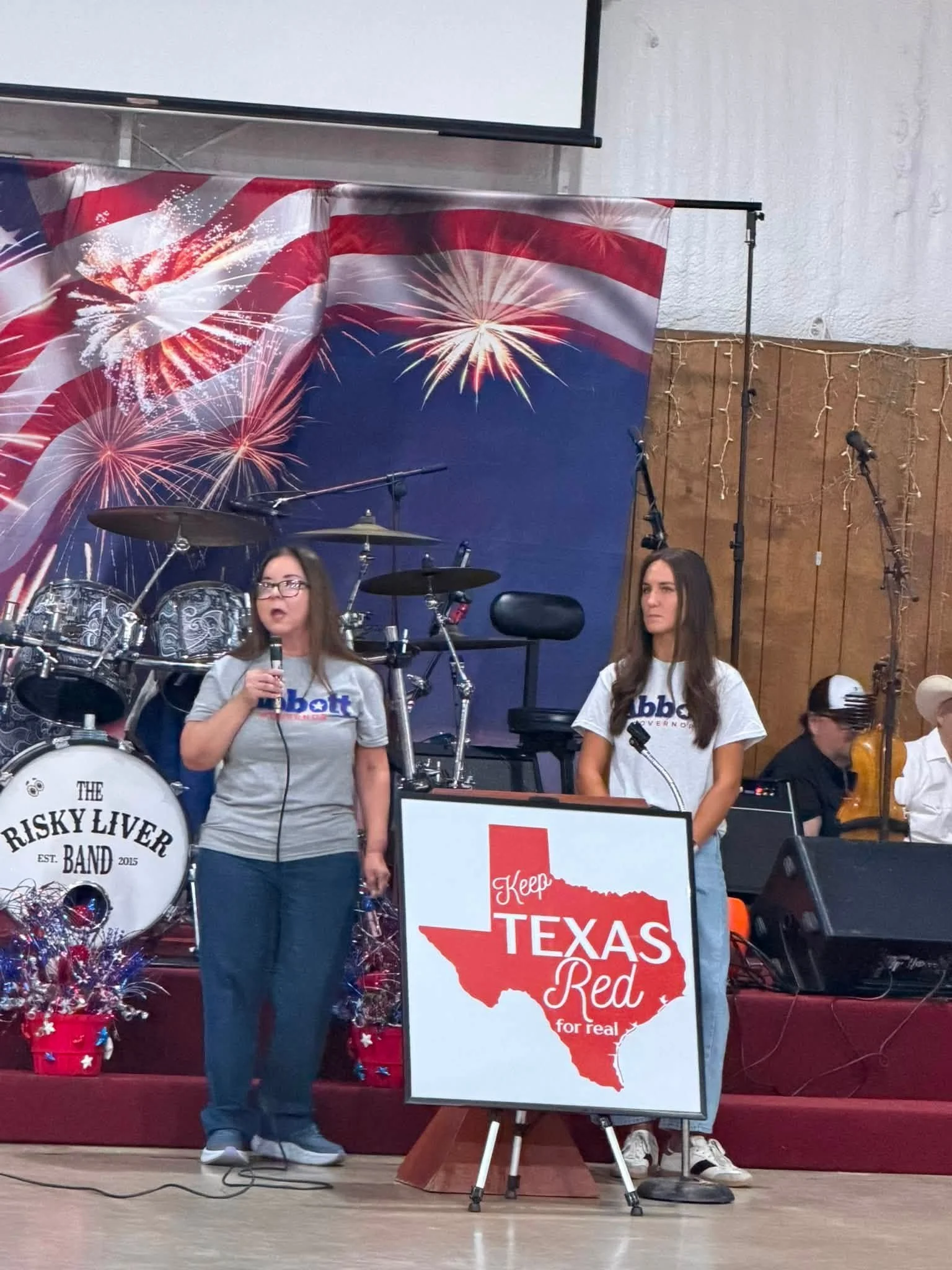 A woman holding a microphone is standing on a stage decorated with patriotic balloons and a banner reading 'Keep Texas Red for real!' The stage backdrop features a large Texas-shaped sign with similar colors and a fireworks display.