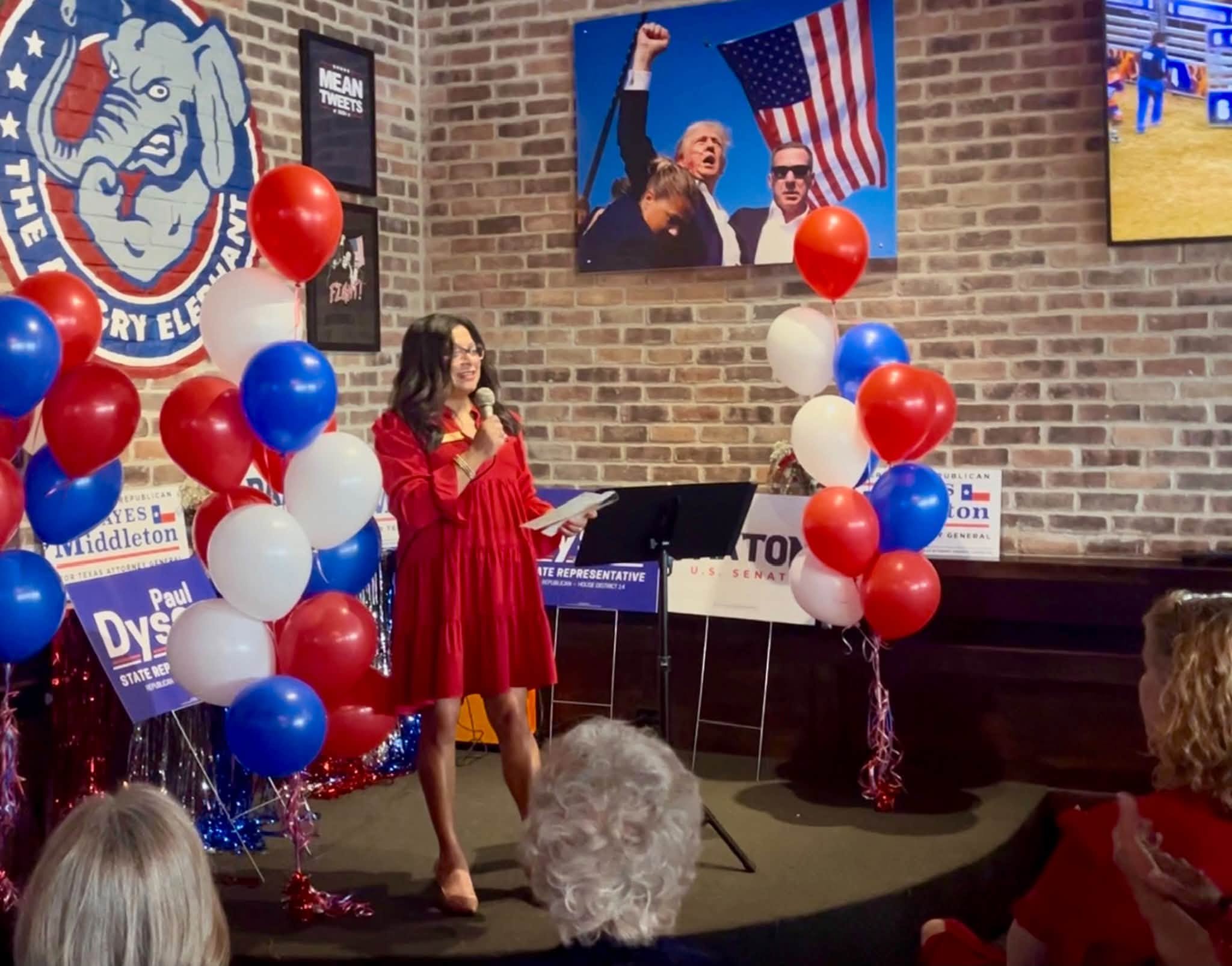 A woman in a red dress stands on stage holding a microphone, speaking to an audience decorated with red, white, and blue balloons and patriotic banners. Behind her, the wall features political posters and a large photograph of a man waving an America