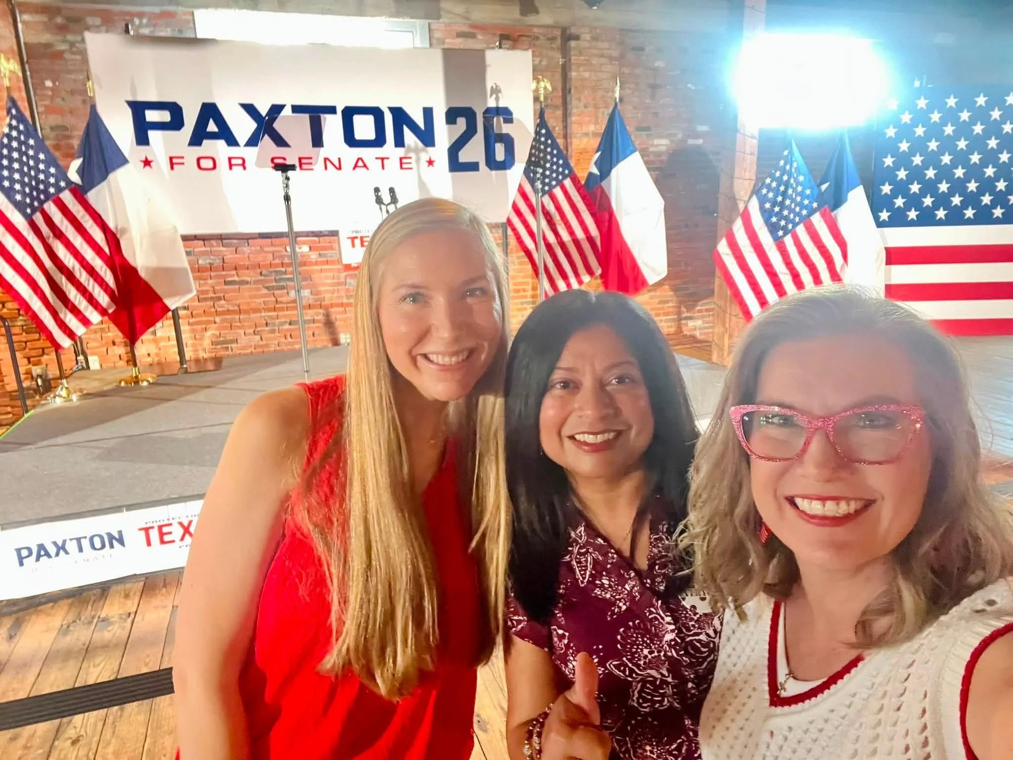 Three women smiling at a political event, with American and Texas flags, and a sign that reads 'PAXTON 26 FOR SENATE' in the background.