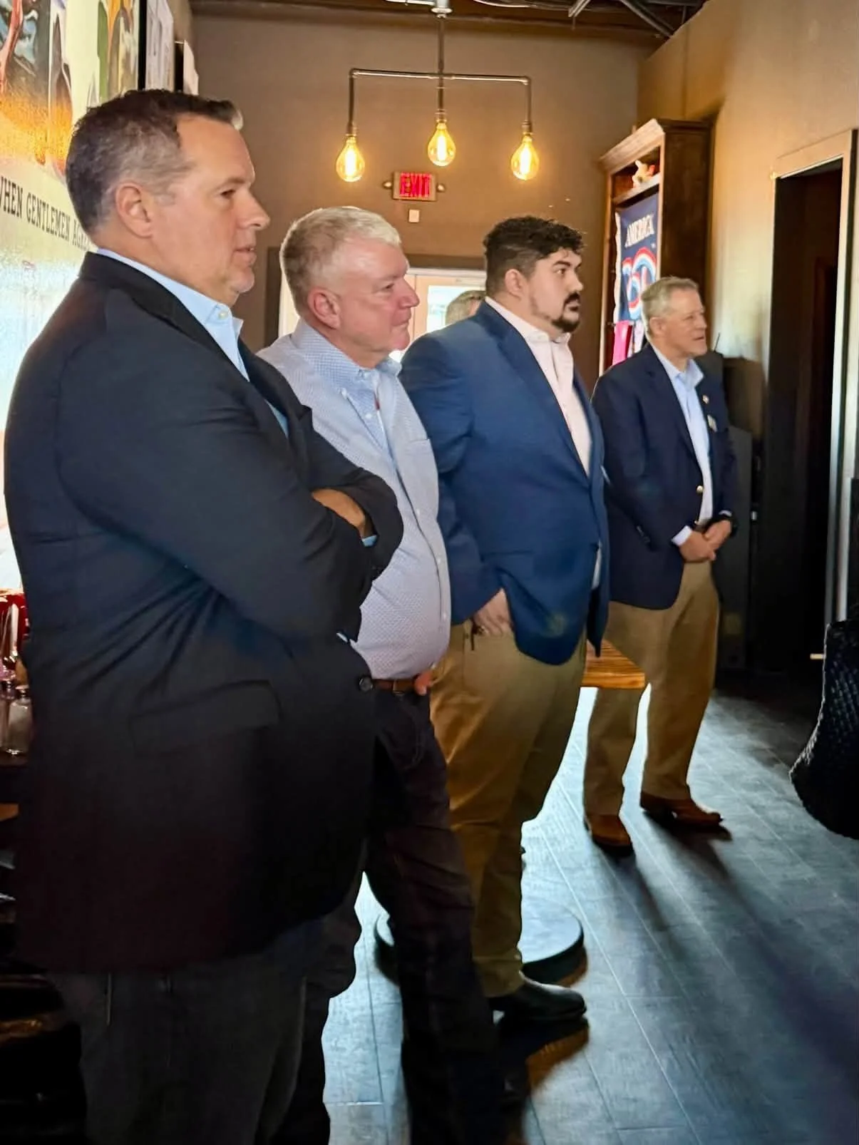 Four men dressed in suits and business casual attire are standing side by side indoors, listening attentively to someone or something outside the frame.