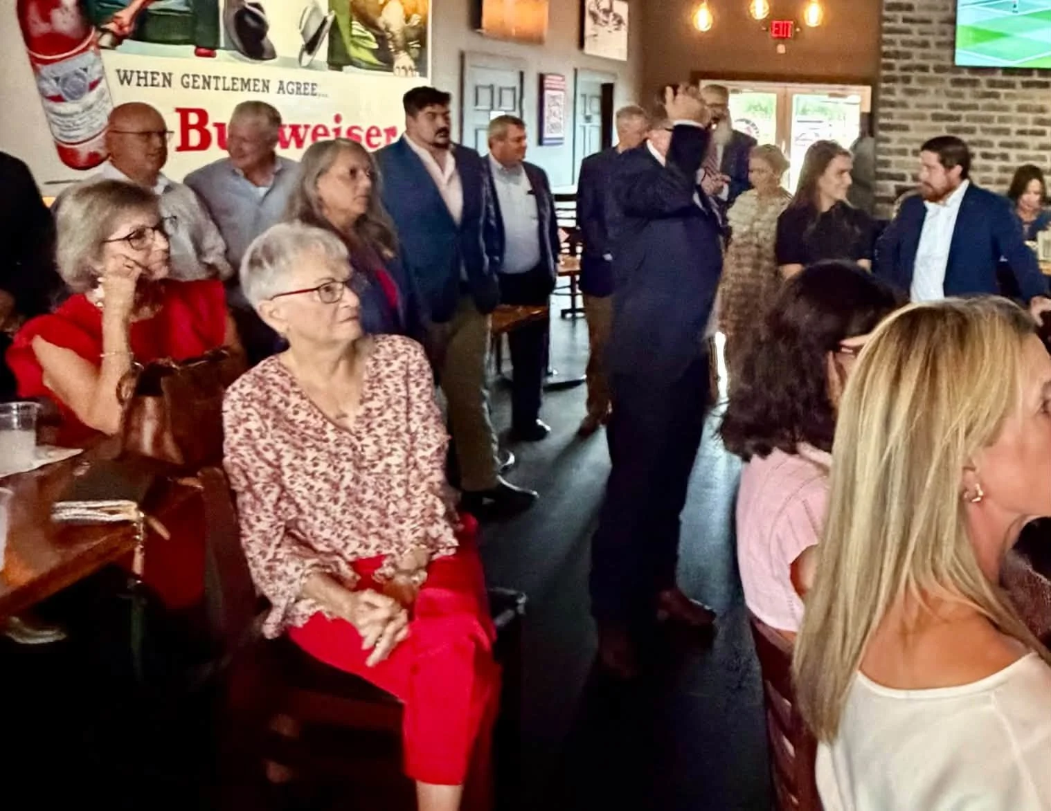 People gathered in a restaurant or bar, some standing and some sitting, with a large Budweiser advertisement on the wall.